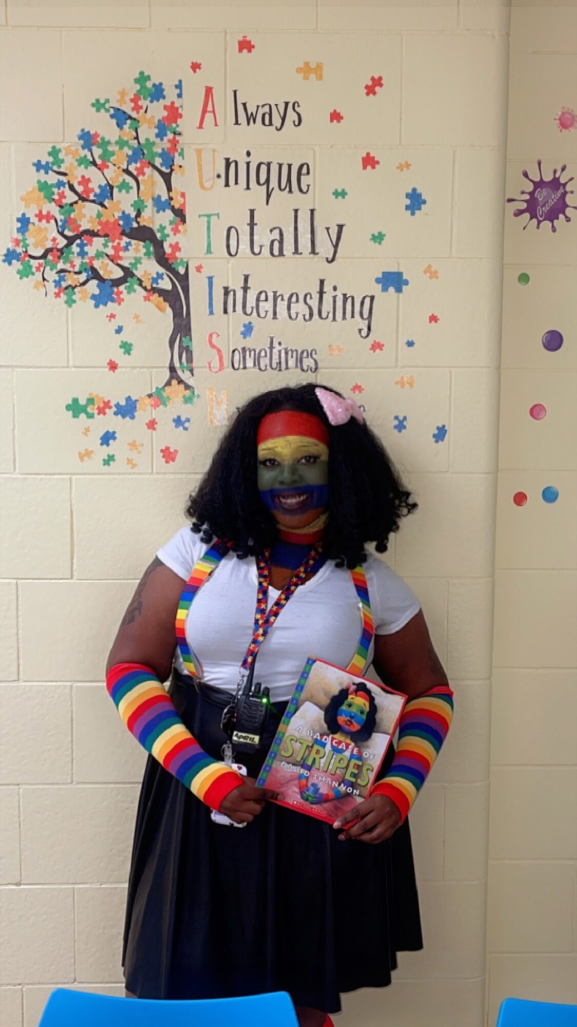 Person wearing rainbow arm warmers and a mask, holding a book, standing in front of a colorful wall with a tree and text.