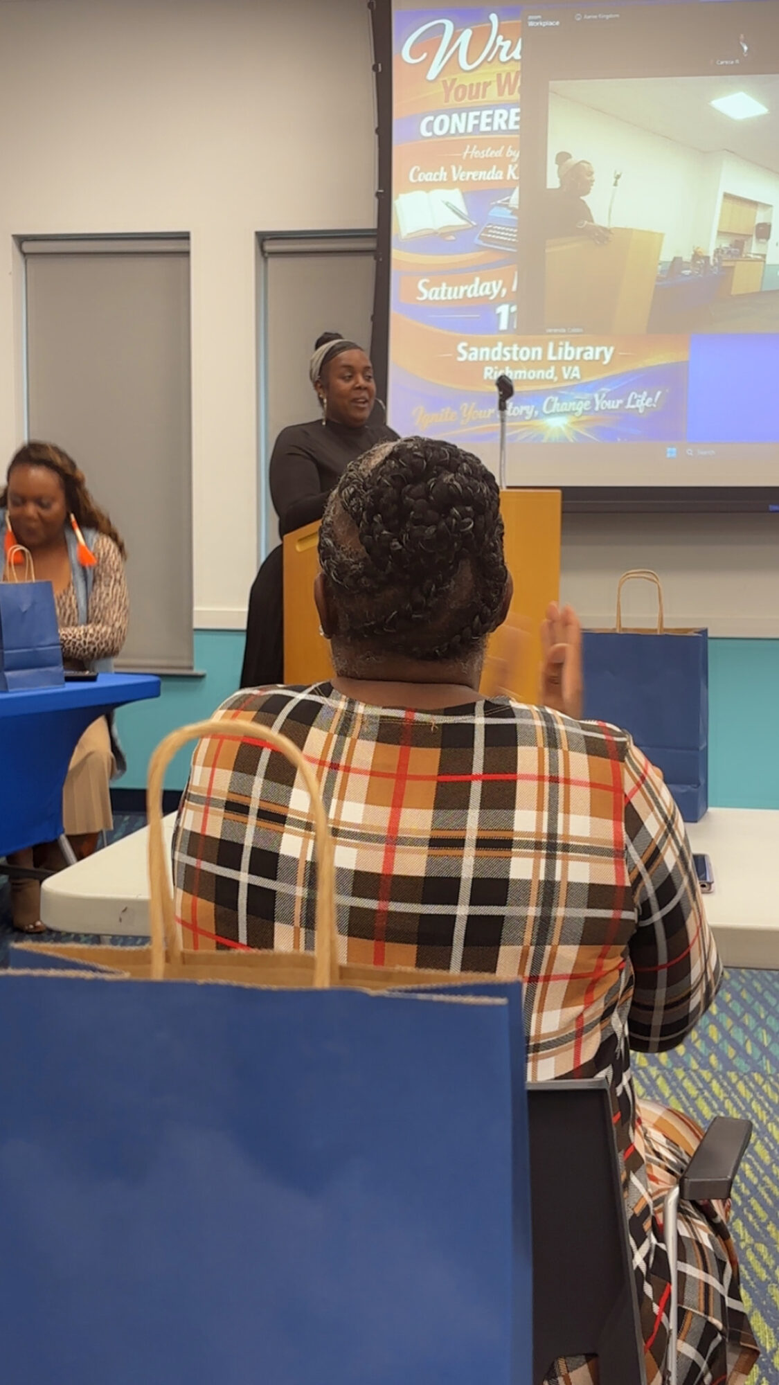 Person speaking at a conference with a projection screen behind, audience seated in a room.
