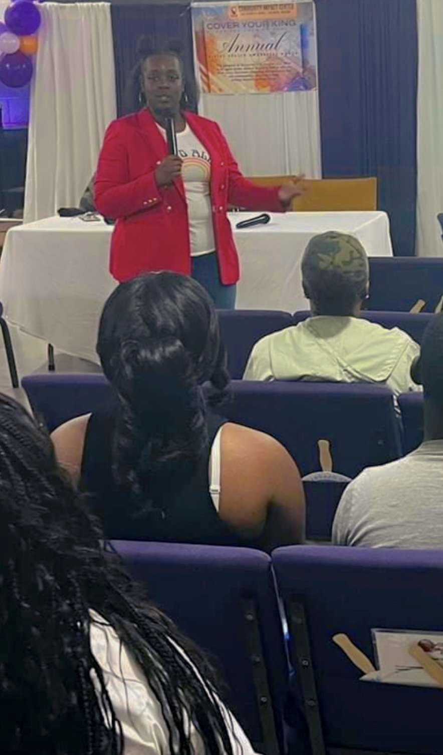 Person in red blazer speaking to an audience in a conference room with a table and banner behind.