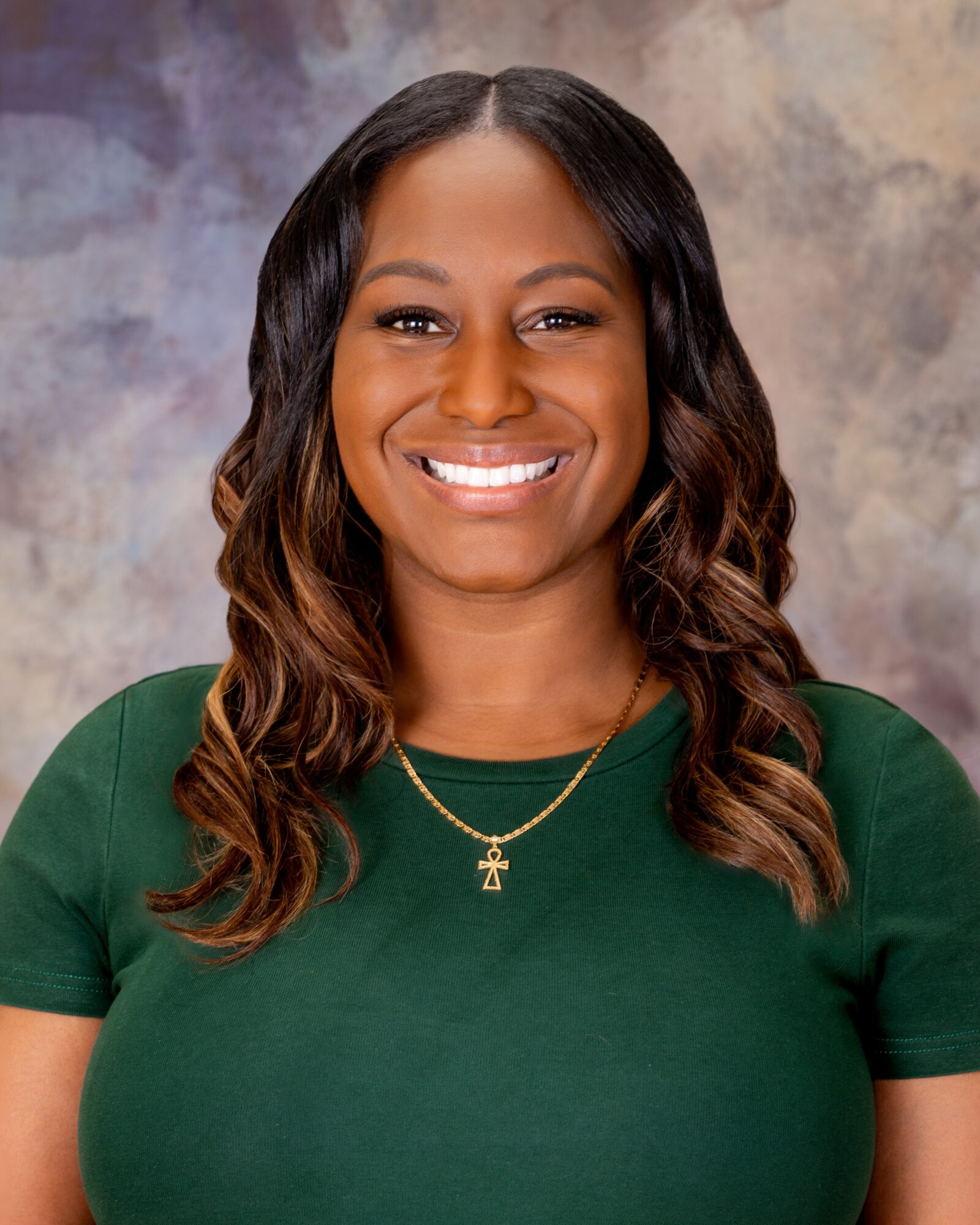 Smiling woman with wavy hair wearing a green top and gold necklace, against a textured background.
