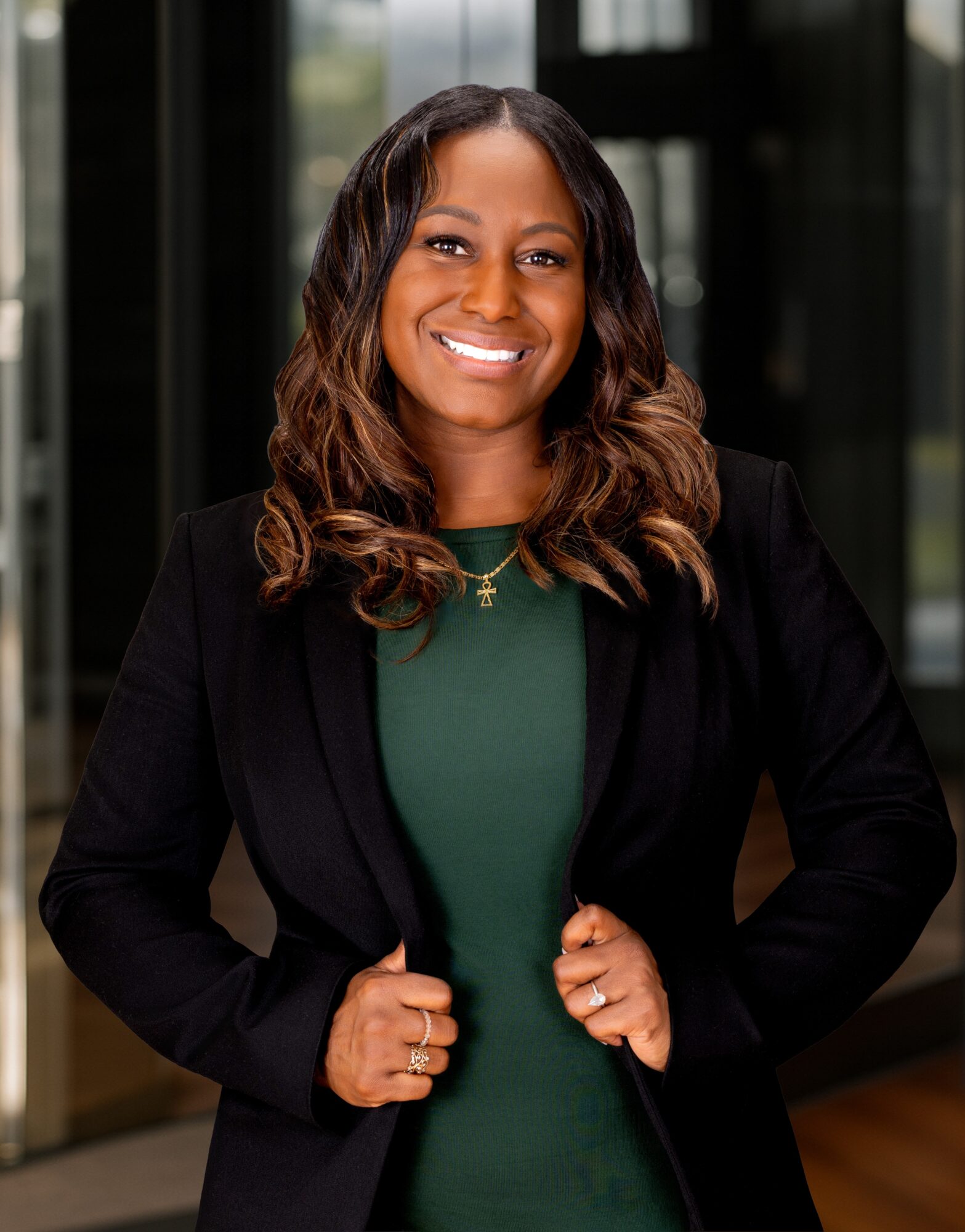 Woman with wavy hair smiling, wearing a black blazer and green top, standing indoors with glass windows behind her.