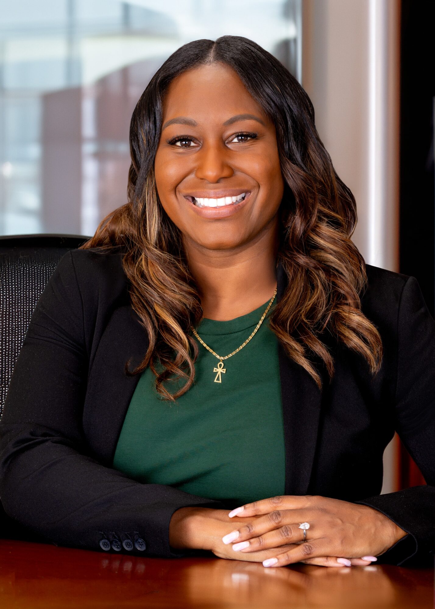 Smiling woman with wavy hair, wearing a green top and black blazer, sitting at a desk in an office.