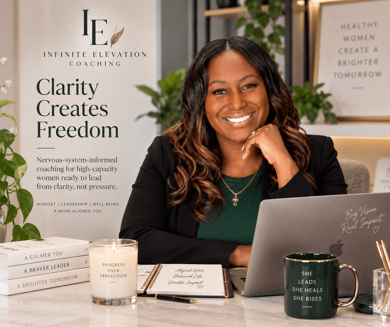 Smiling woman with long dark hair sitting at a desk with a laptop, books, and a glass of water, in an office setting.