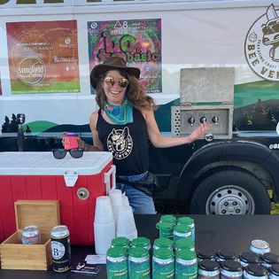 Woman with sunglasses and hat standing behind a table with drinks and a cooler, outdoor event setting.