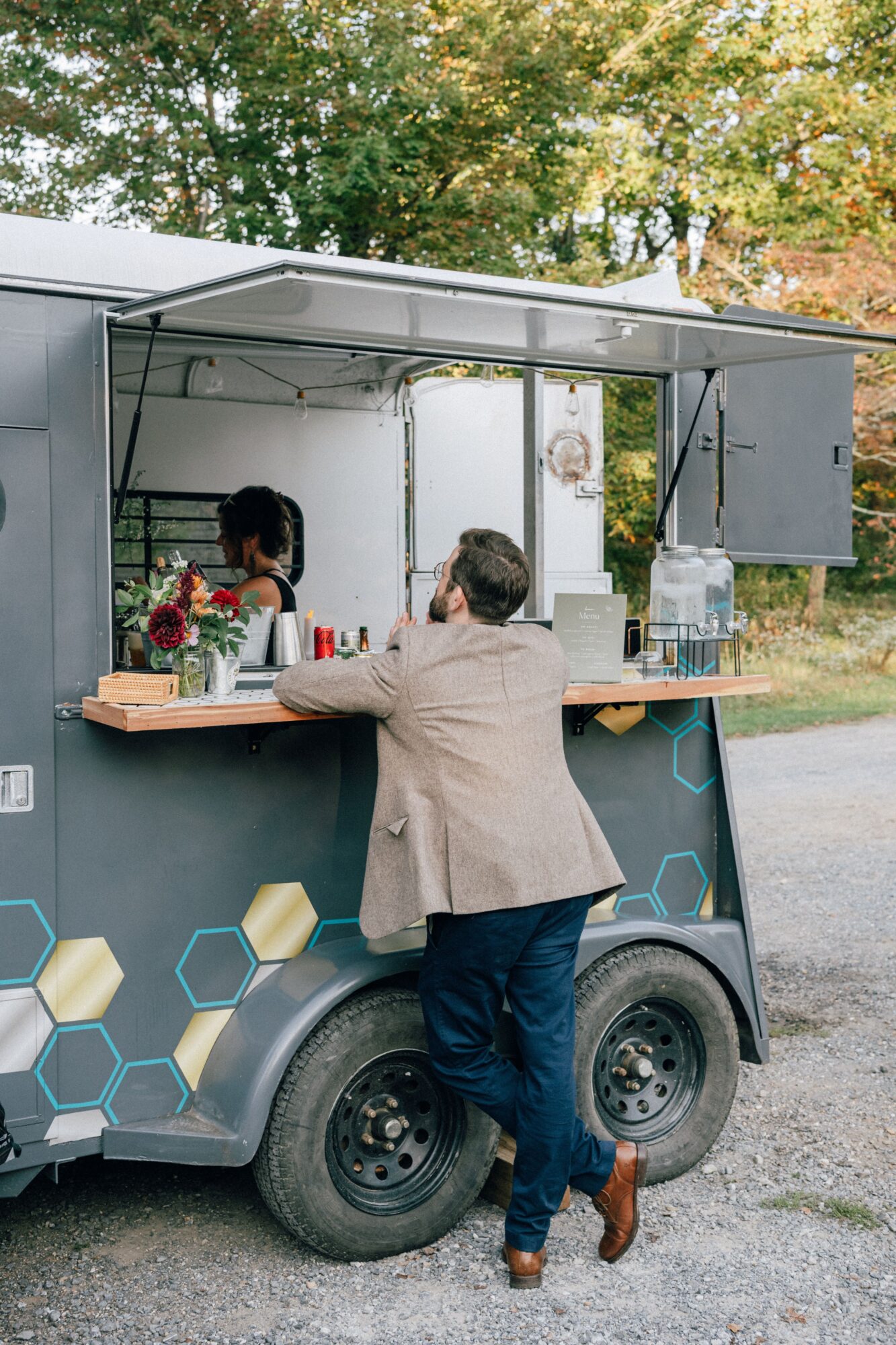 Person ordering at a mobile food truck with a woman inside, outdoor setting with trees, and a wooden counter.
