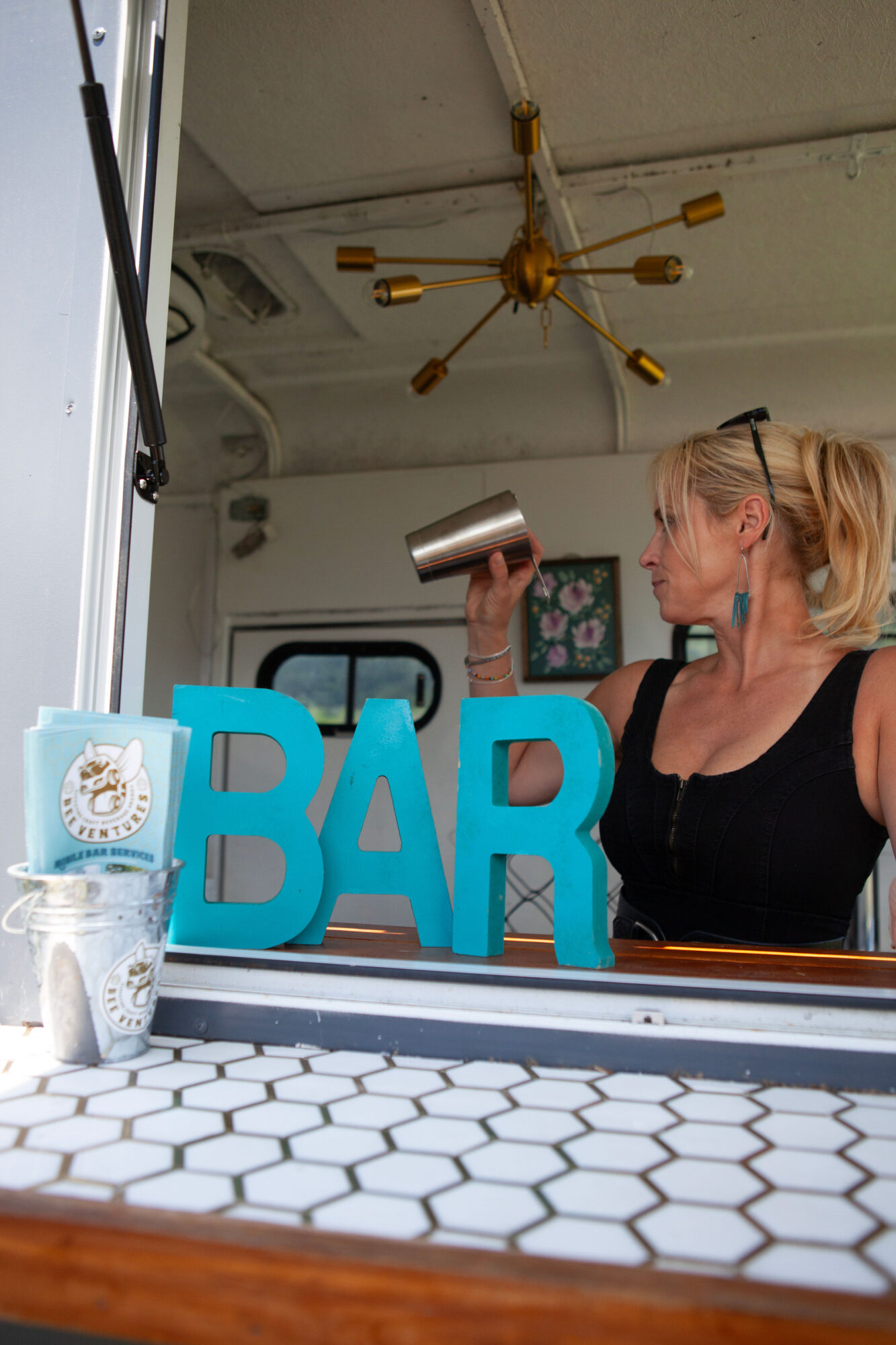 A woman with blonde hair in a ponytail drinks from a metal cup behind a bar with large blue letters spelling 'BAR'.