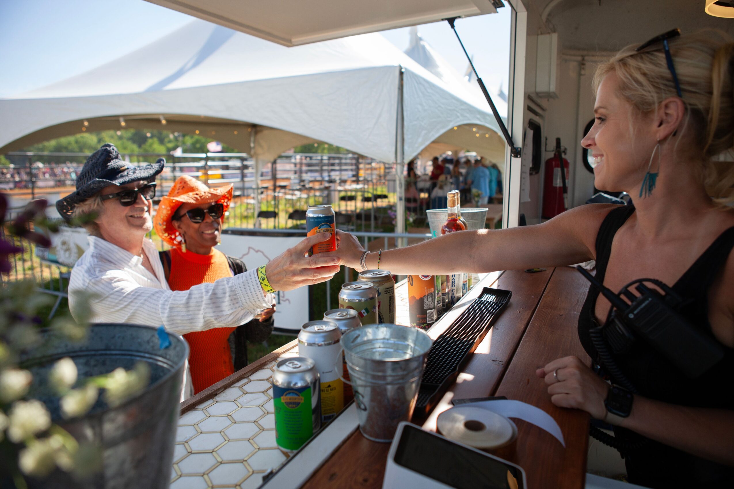 People at a booth exchanging drinks, with a woman smiling and two children wearing hats, outdoors under a tent.
