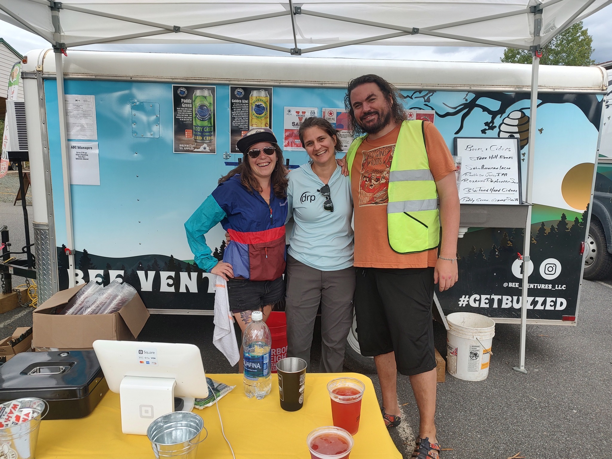 Three people standing under a tent, smiling, with posters and a whiteboard behind them, outdoor setting.