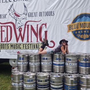 Person lying on top of beer kegs at outdoor music festival, with a banner in the background displaying event and brewery logos.