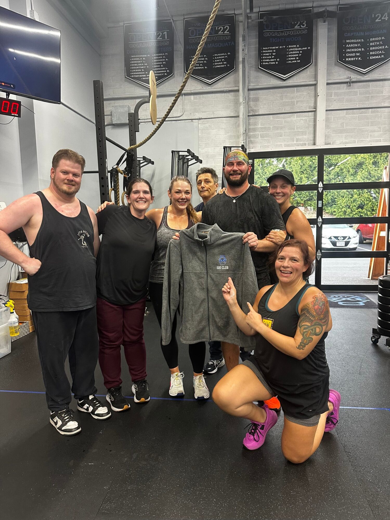 Group of six people in gym, one holding a gray hoodie, others smiling, some with tattoos, gym equipment visible in background.