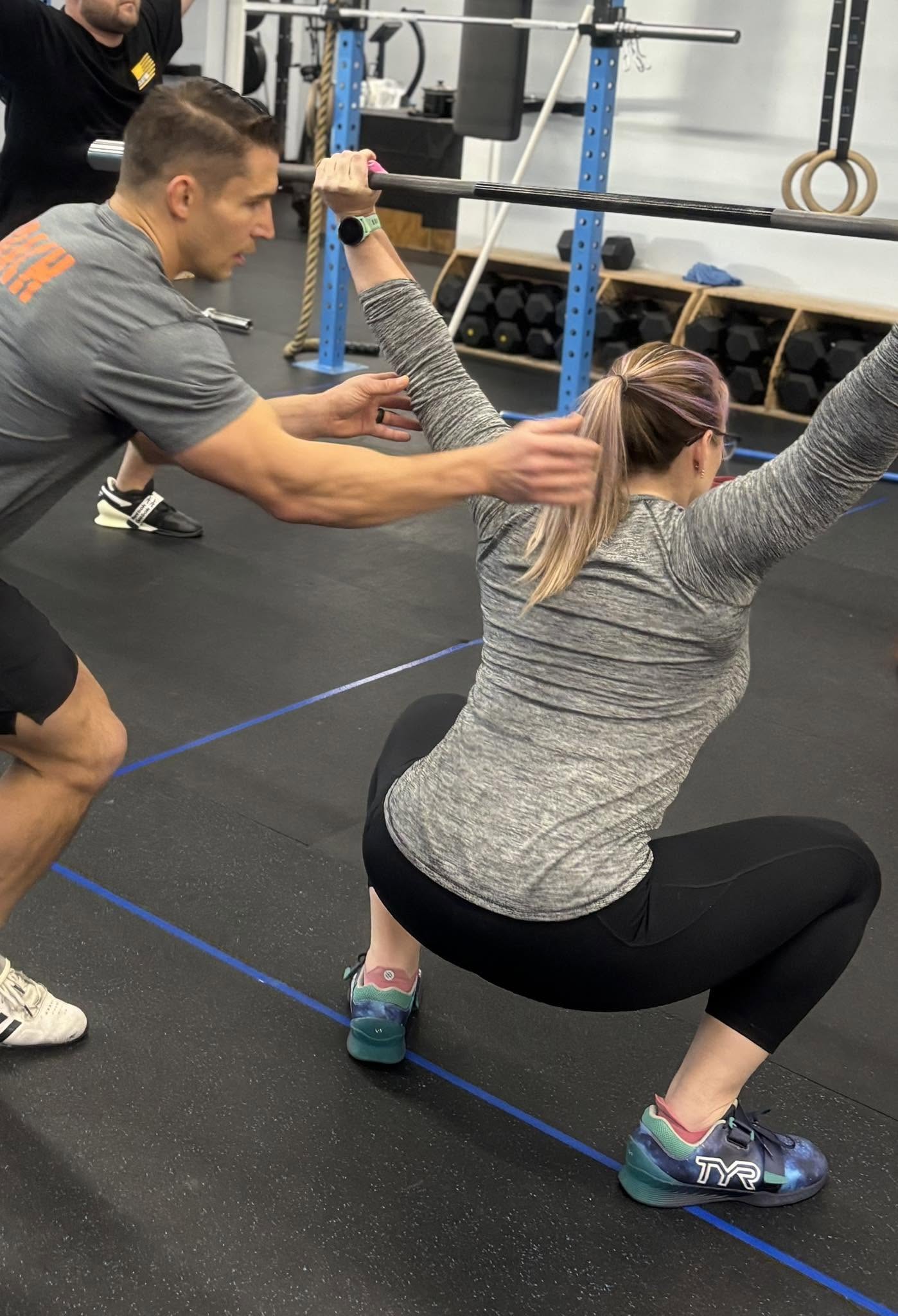 Personal trainer assists woman during squat exercise in gym, with gym equipment in background.