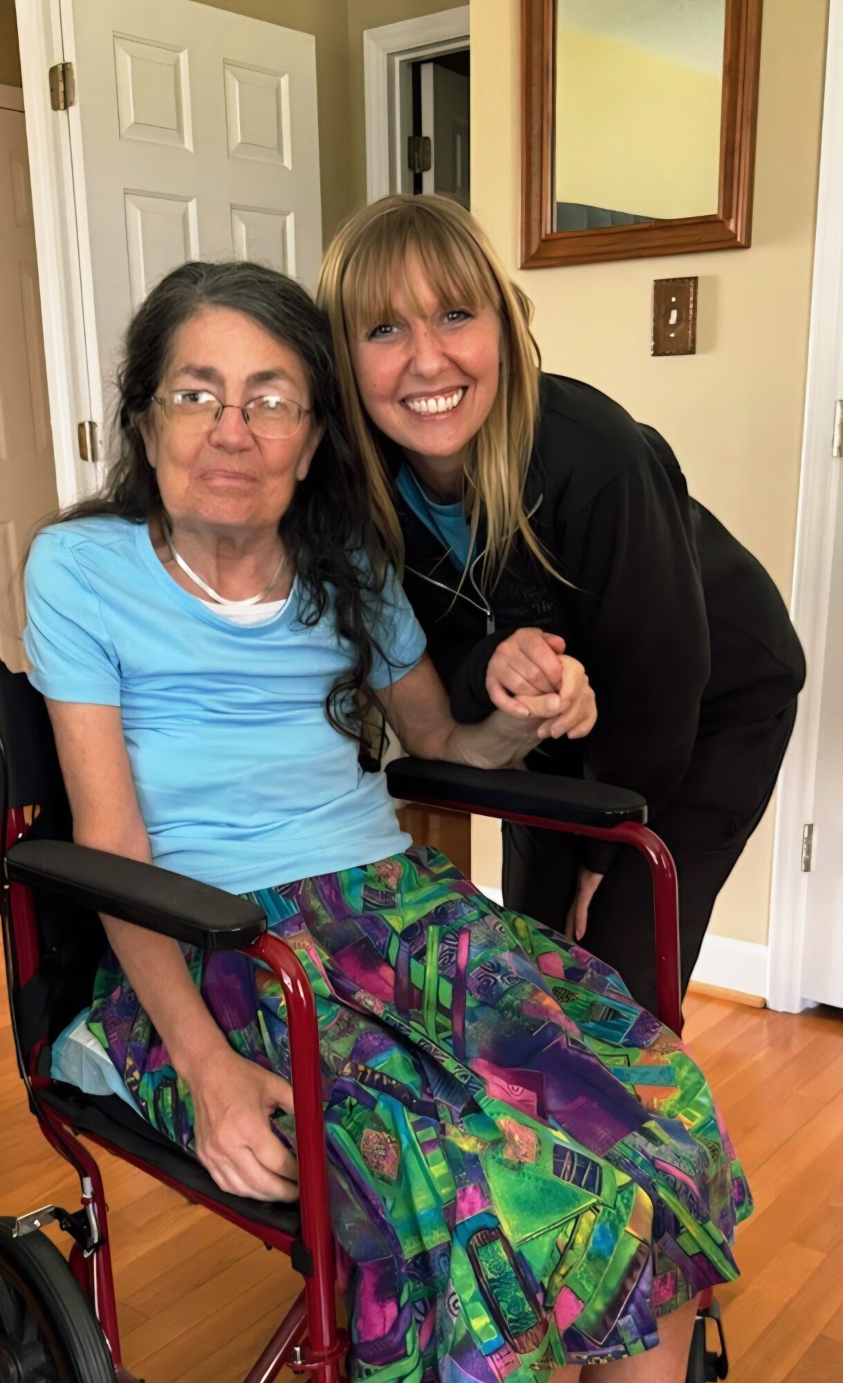 Two women smiling indoors, one sitting in a wheelchair and the other standing beside her, both looking at the camera.
