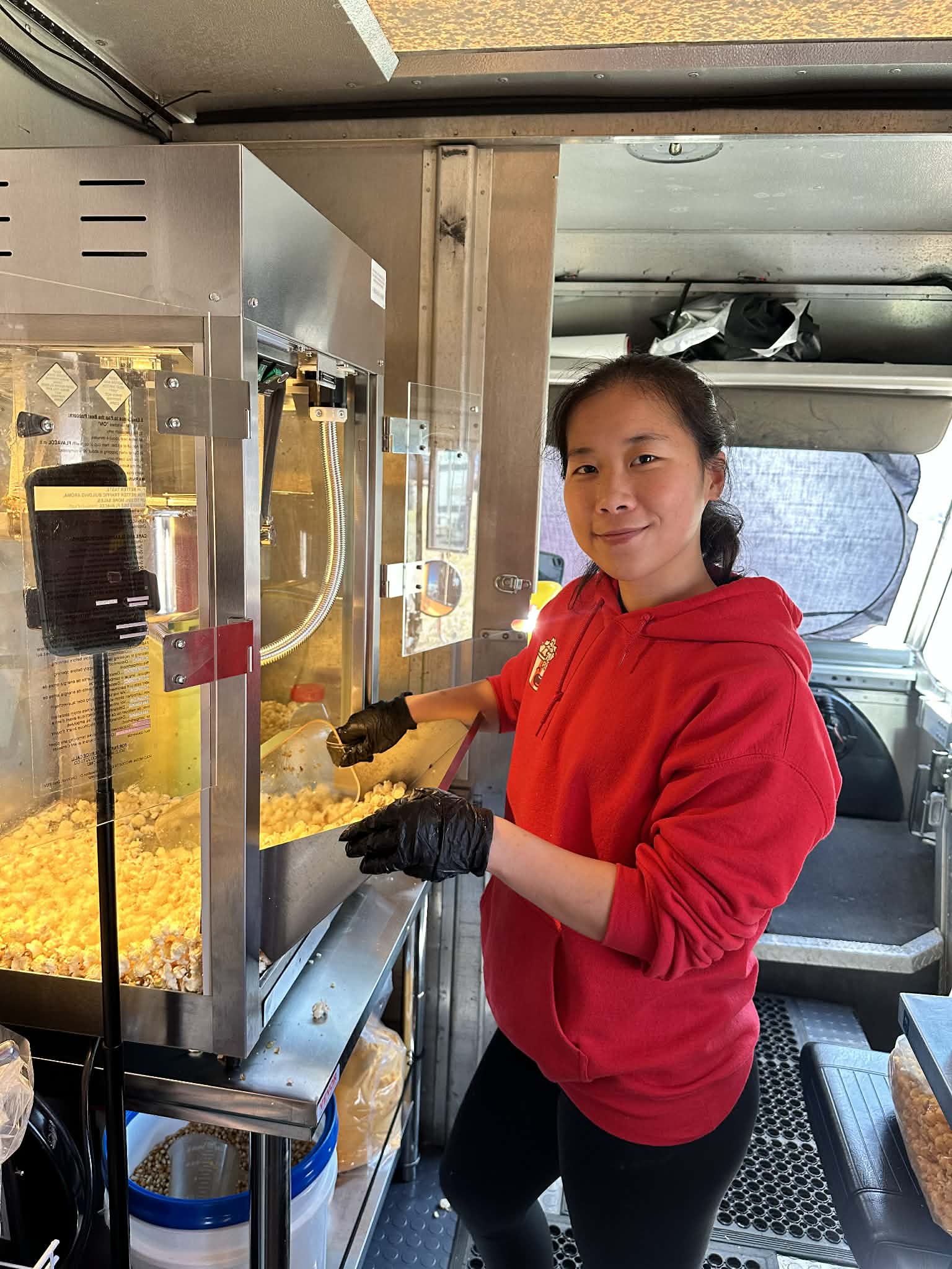 Young woman in red hoodie and black gloves operating popcorn machine indoors.