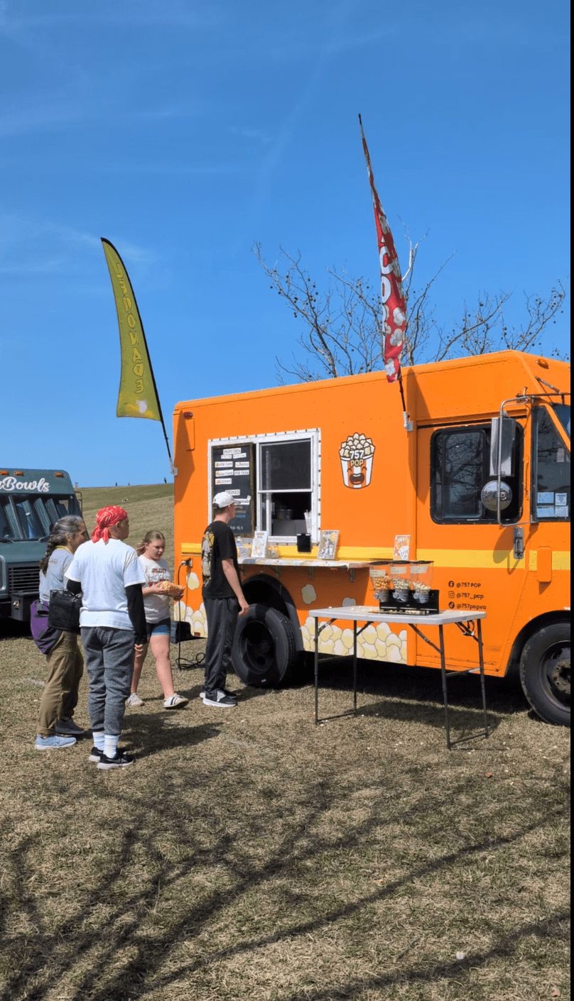 People gather around an orange food truck with flags on a sunny day, with a clear blue sky and grassy area.