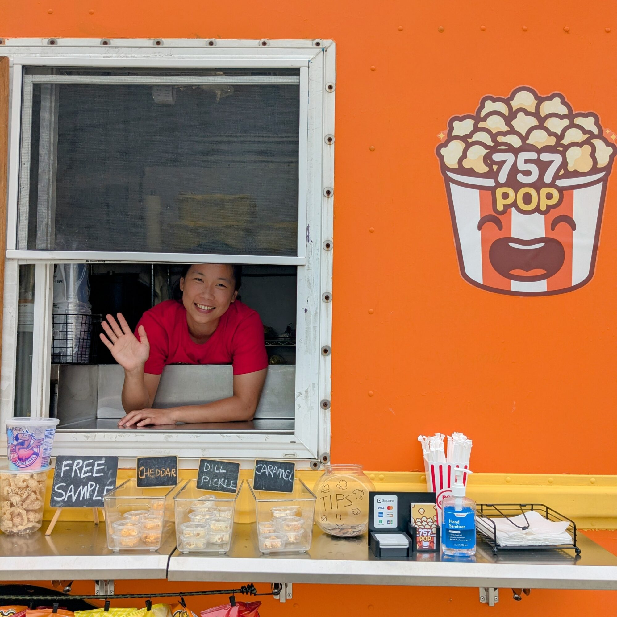 Girl smiling and waving at a popcorn stand window with a popcorn logo on an orange background.
