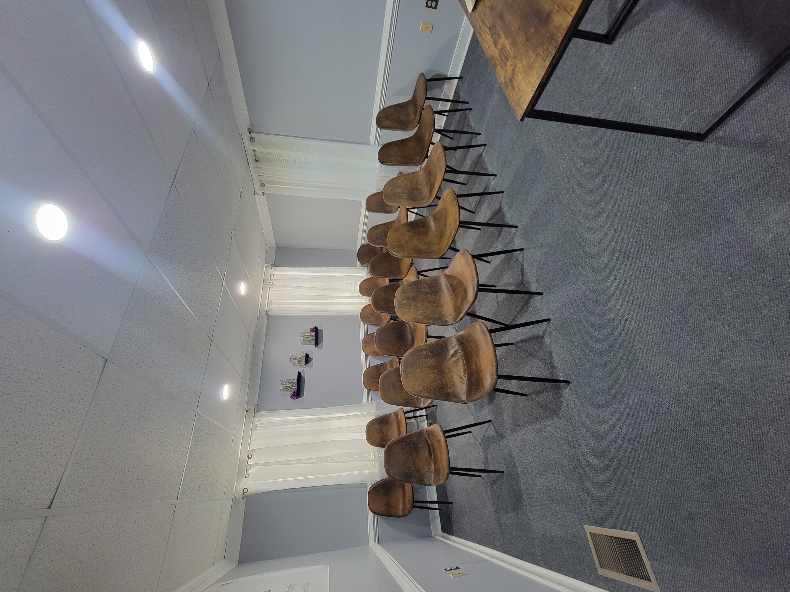 Row of wooden chairs with black metal legs in a room with white walls and ceiling, gray carpeted floor, and a table.