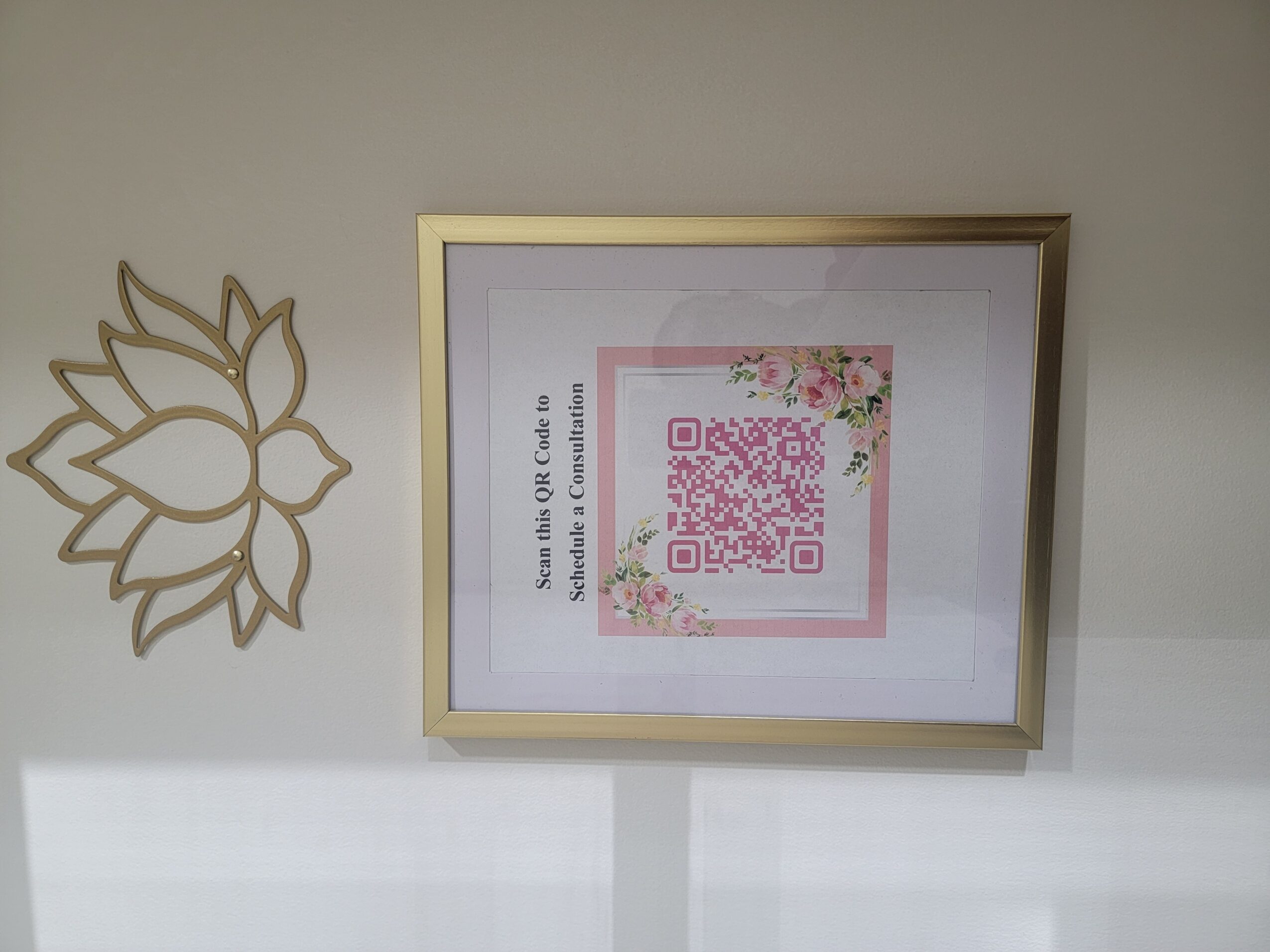 Framed poster with a pink QR code and floral design, hanging on a wall, with a decorative metal flower on the left.