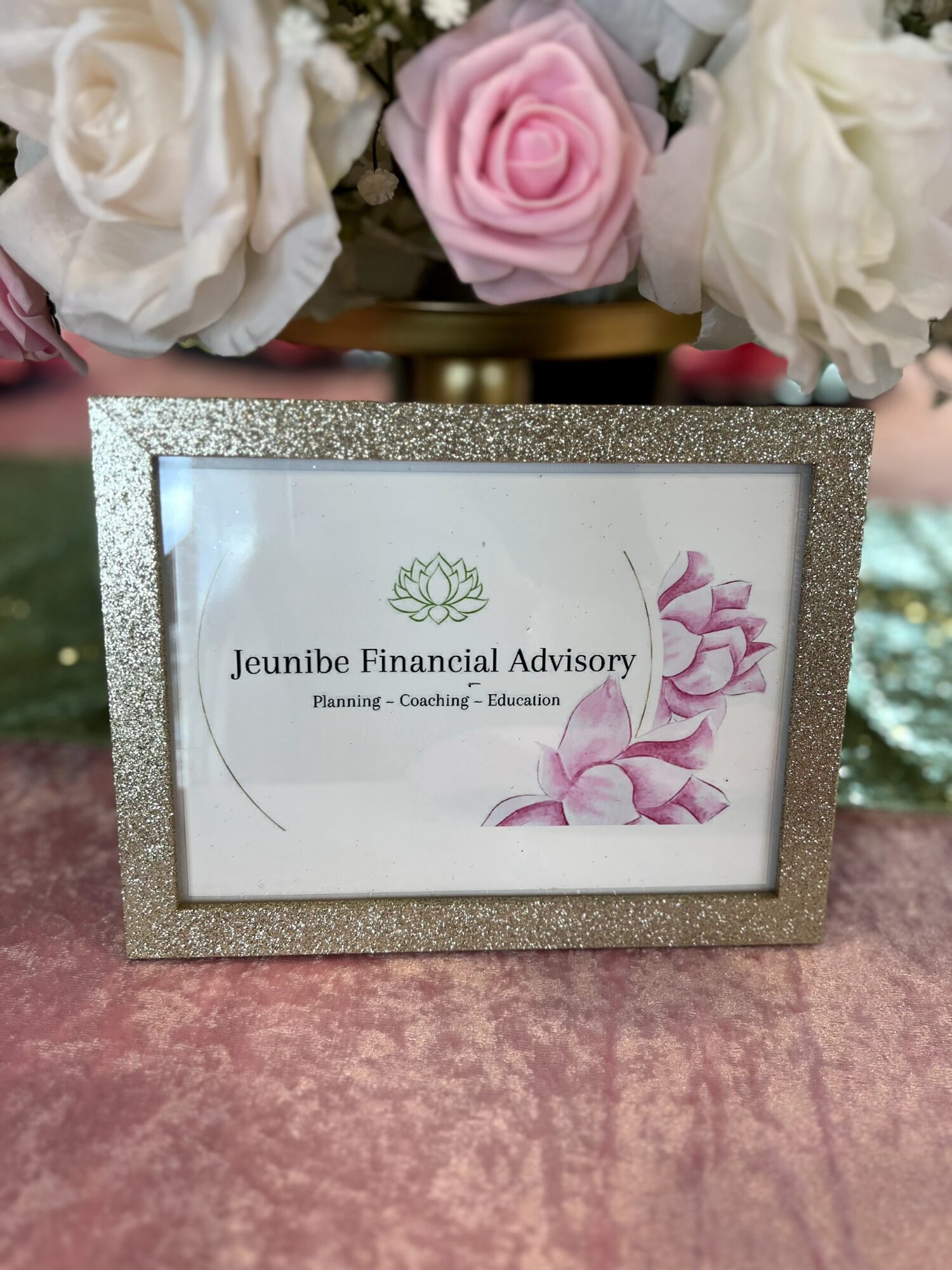 Framed certificate with glittery border, displaying Jeunibe Financial Advisory logo and text, with pink floral design, on a pink surface.