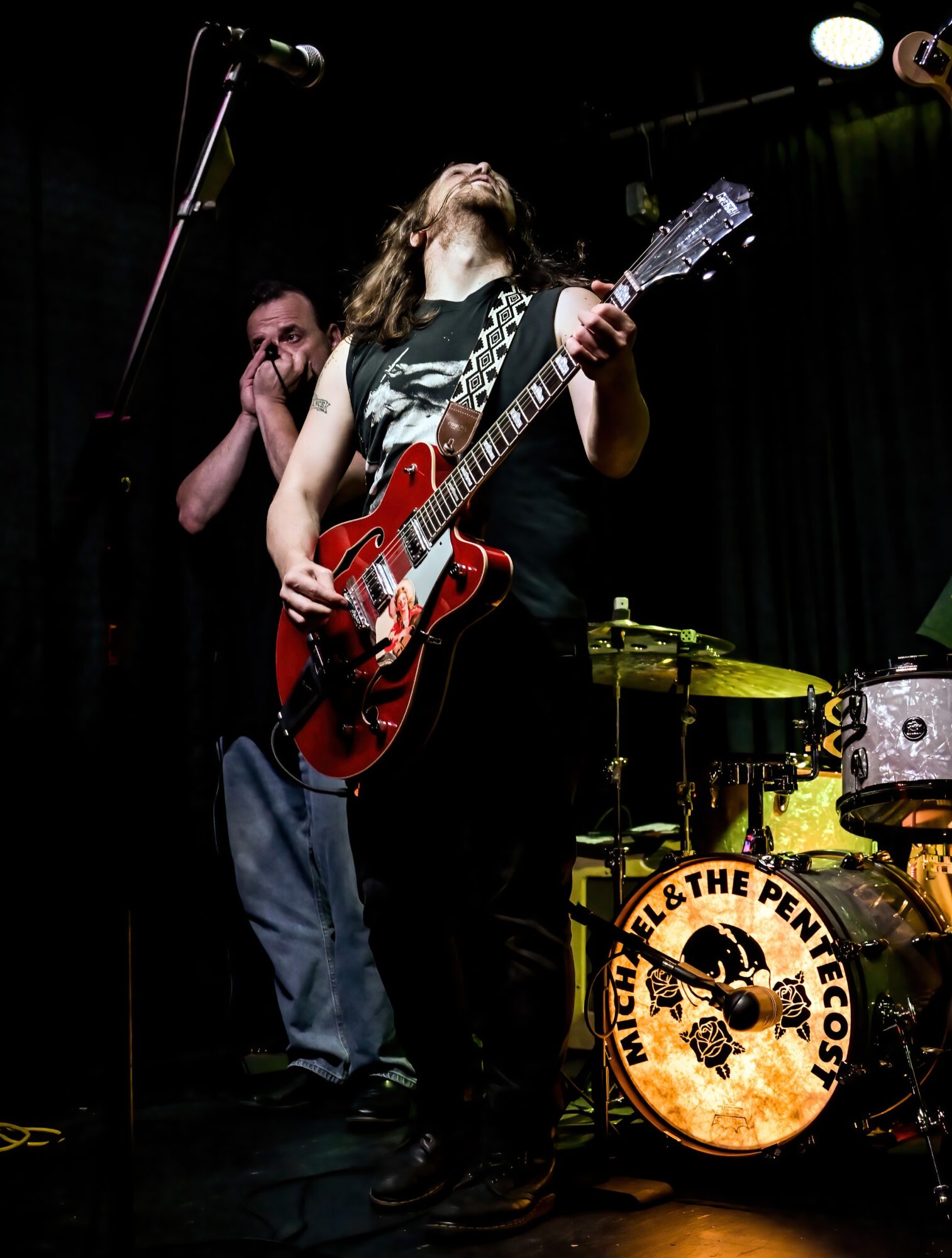 Musician playing guitar on stage with a drummer in the background, dark stage with black curtains, and a drum set with a logo.