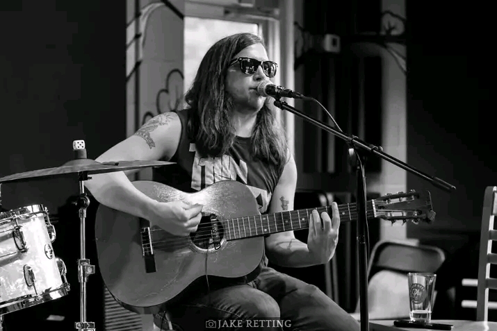 Woman with sunglasses playing guitar and singing into microphone on stage, with drum set and chair nearby, in black and white.