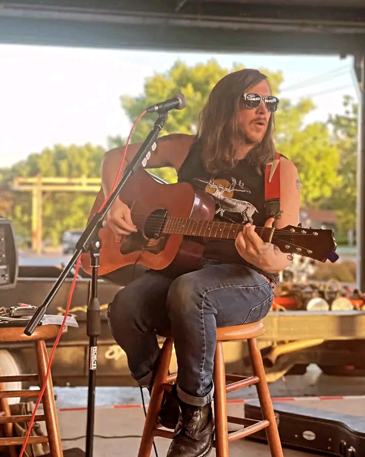 Person with long hair, sunglasses, and a red sleeveless shirt plays guitar and sings into a microphone outdoors.