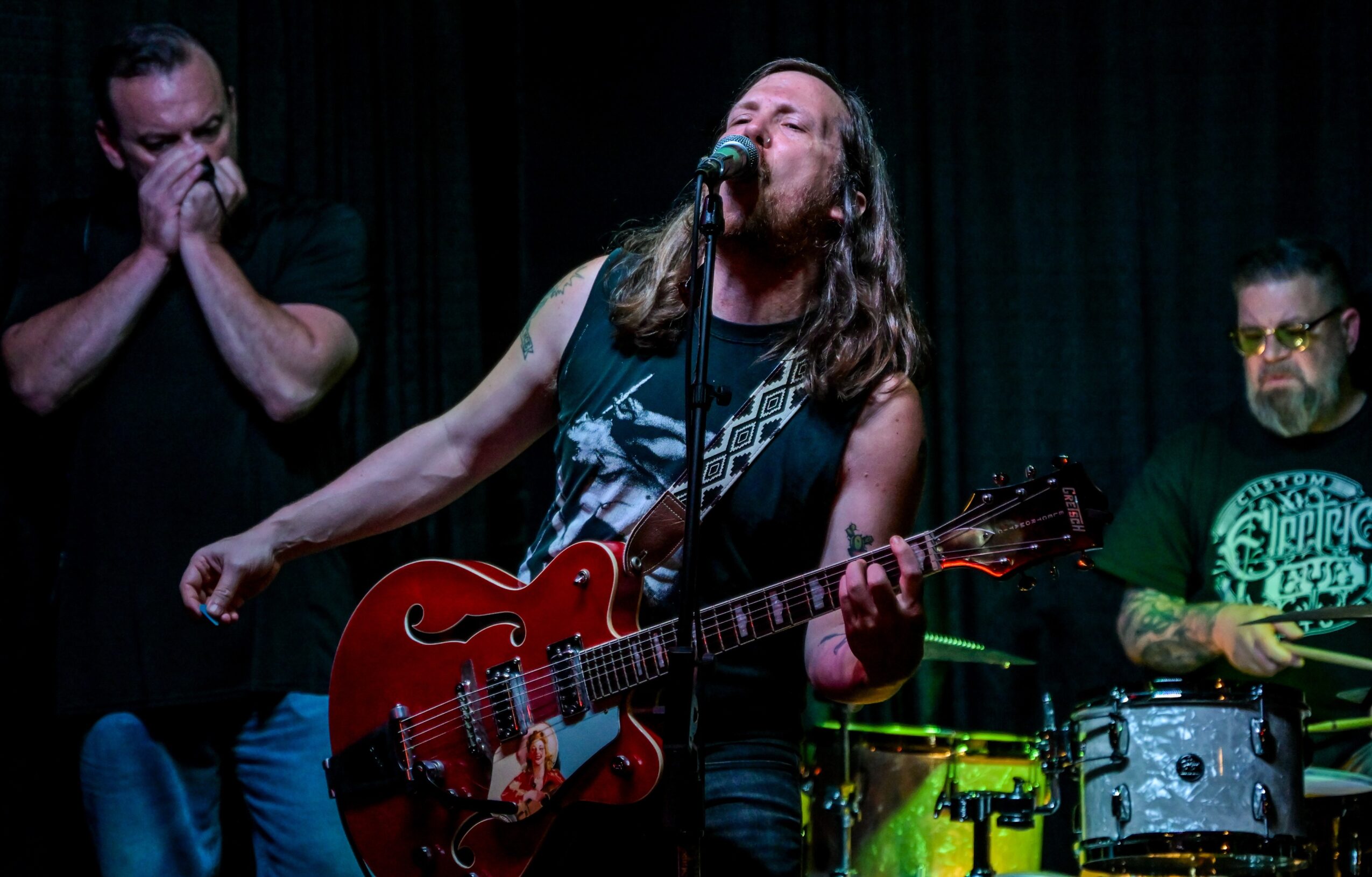 Three musicians perform on stage, one singing with guitar, others playing percussion, dark background, stage lighting.