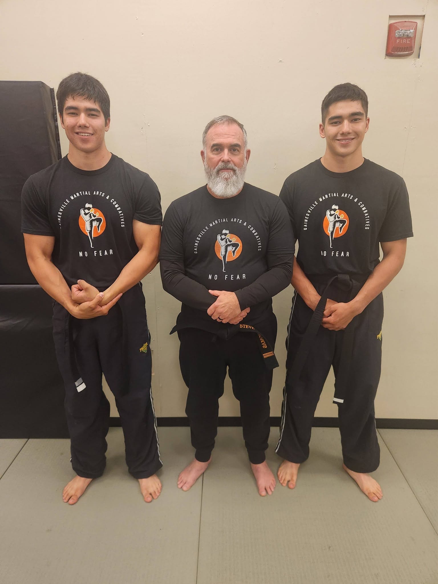 Three people standing barefoot against a wall, two young men and an older man in the middle, all wearing black shirts with a logo.