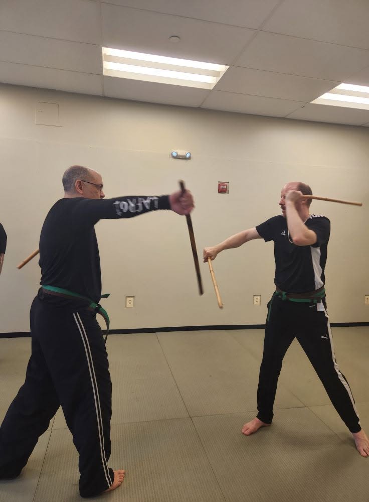 Two people practicing martial arts with wooden sticks in a room, one person blocking the other's strike, both barefoot.
