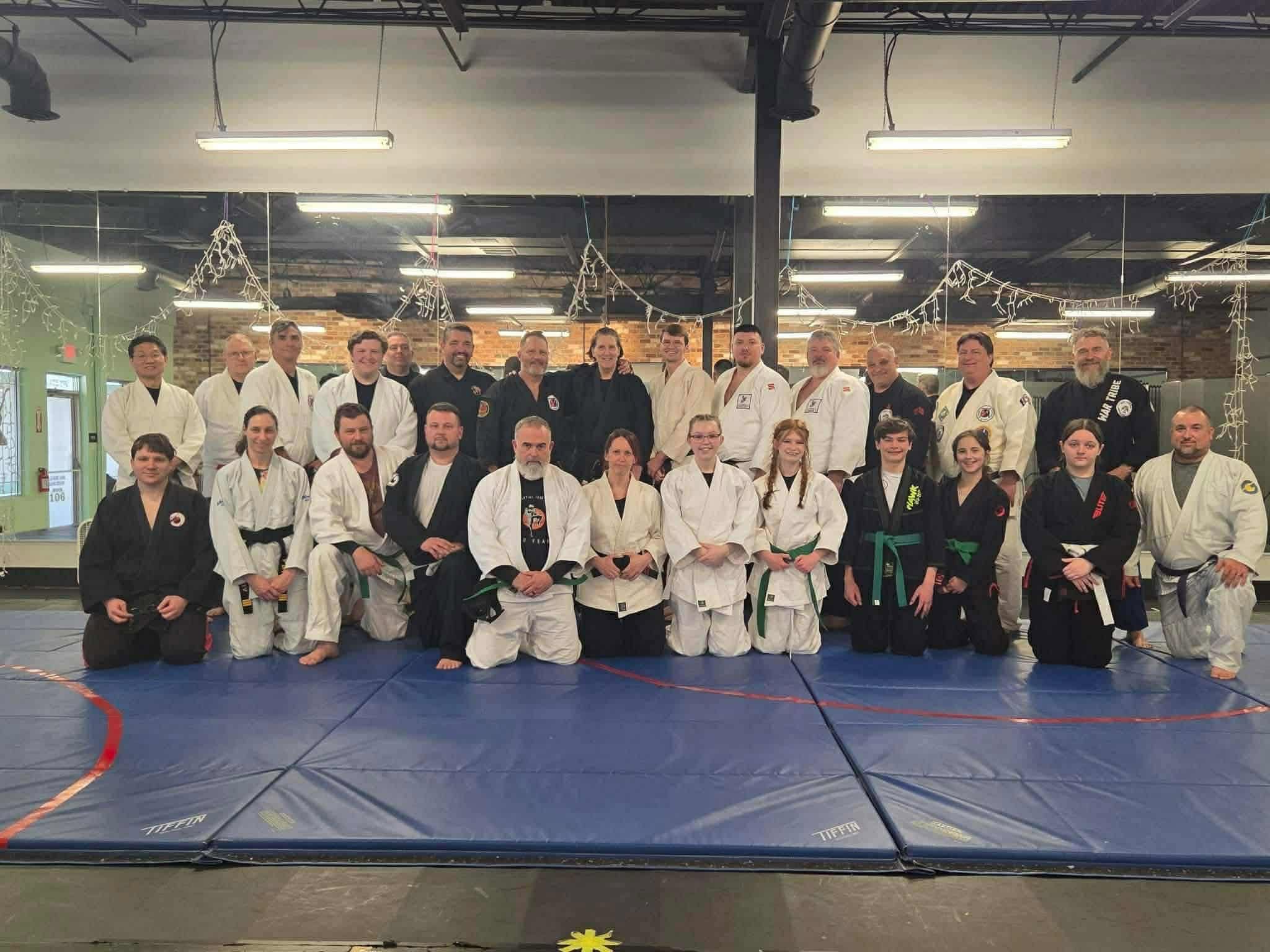 Group of people in martial arts uniforms and gis, posing on a blue mat in a dojo or gym, some kneeling and some standing.