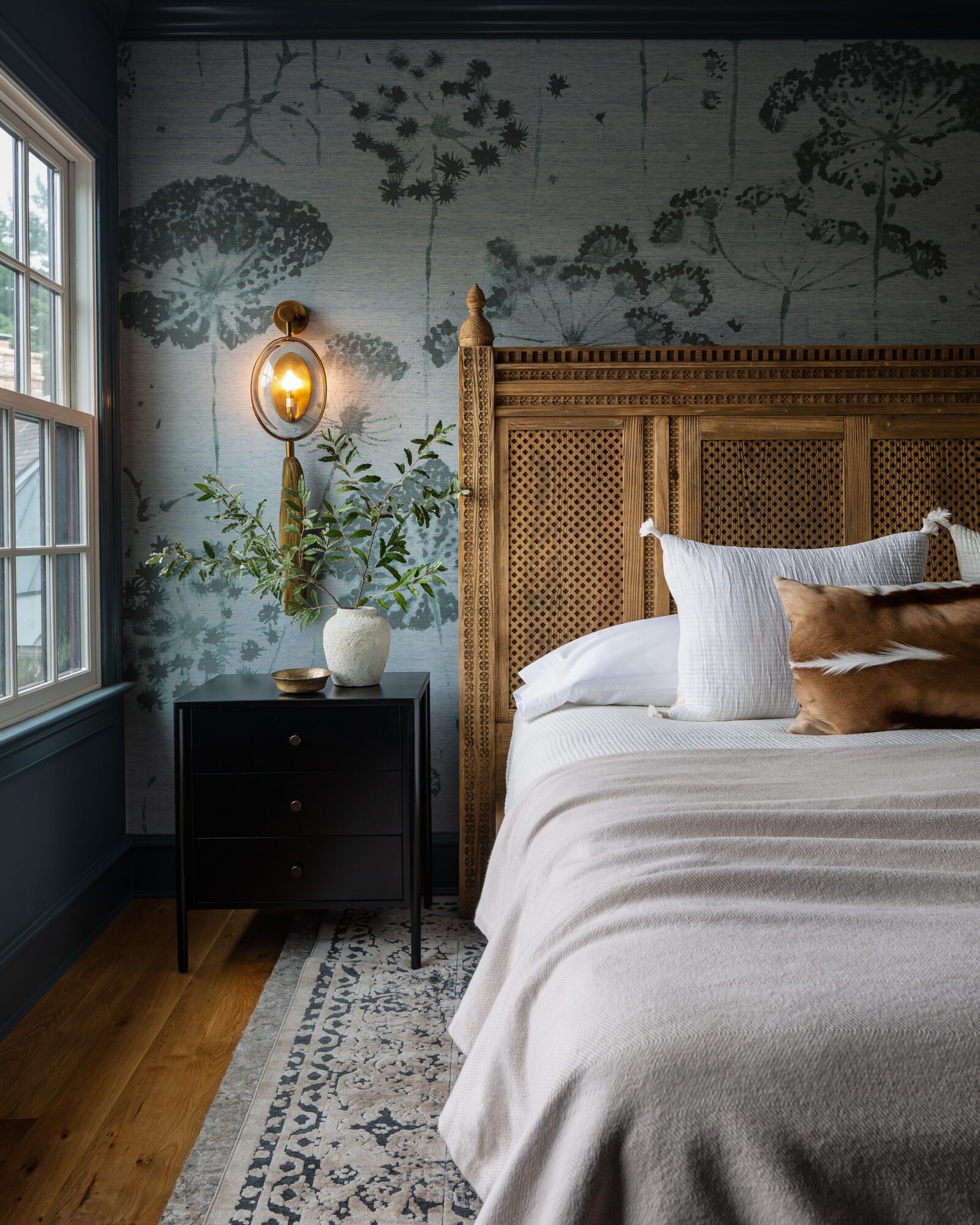 Bedroom corner with a bed, wooden headboard, nightstand, and a wall with tree-patterned wallpaper, illuminated by a wall-mounted lamp.