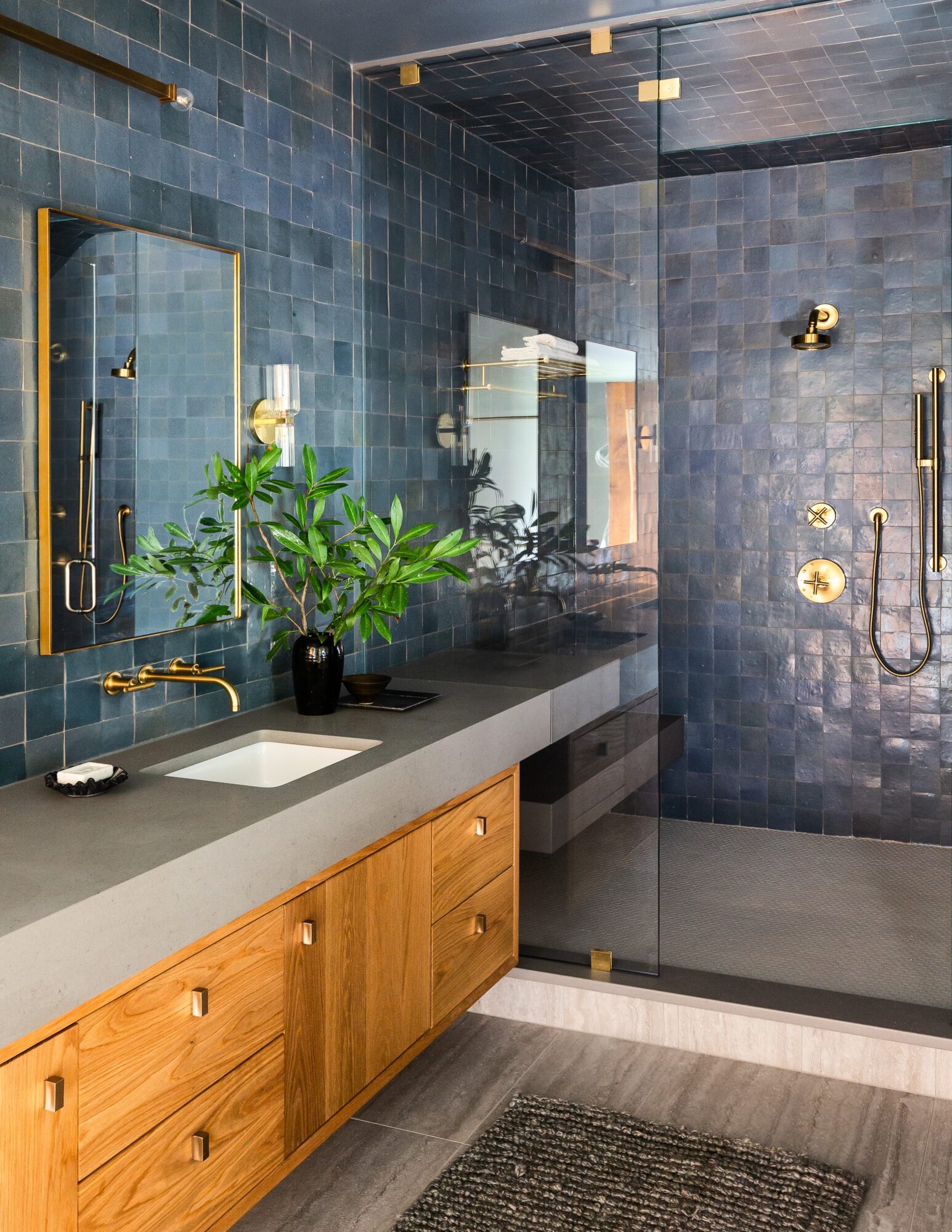 Bathroom with blue tiled walls, wooden vanity, mirror, plant, and shower area with gold fixtures.