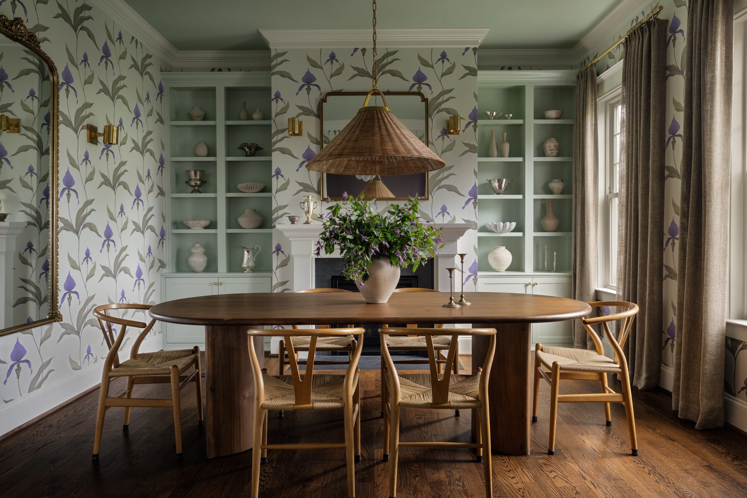 Dining room with a round wooden table, six chairs, floral wallpaper, built-in shelves, and large windows with curtains.