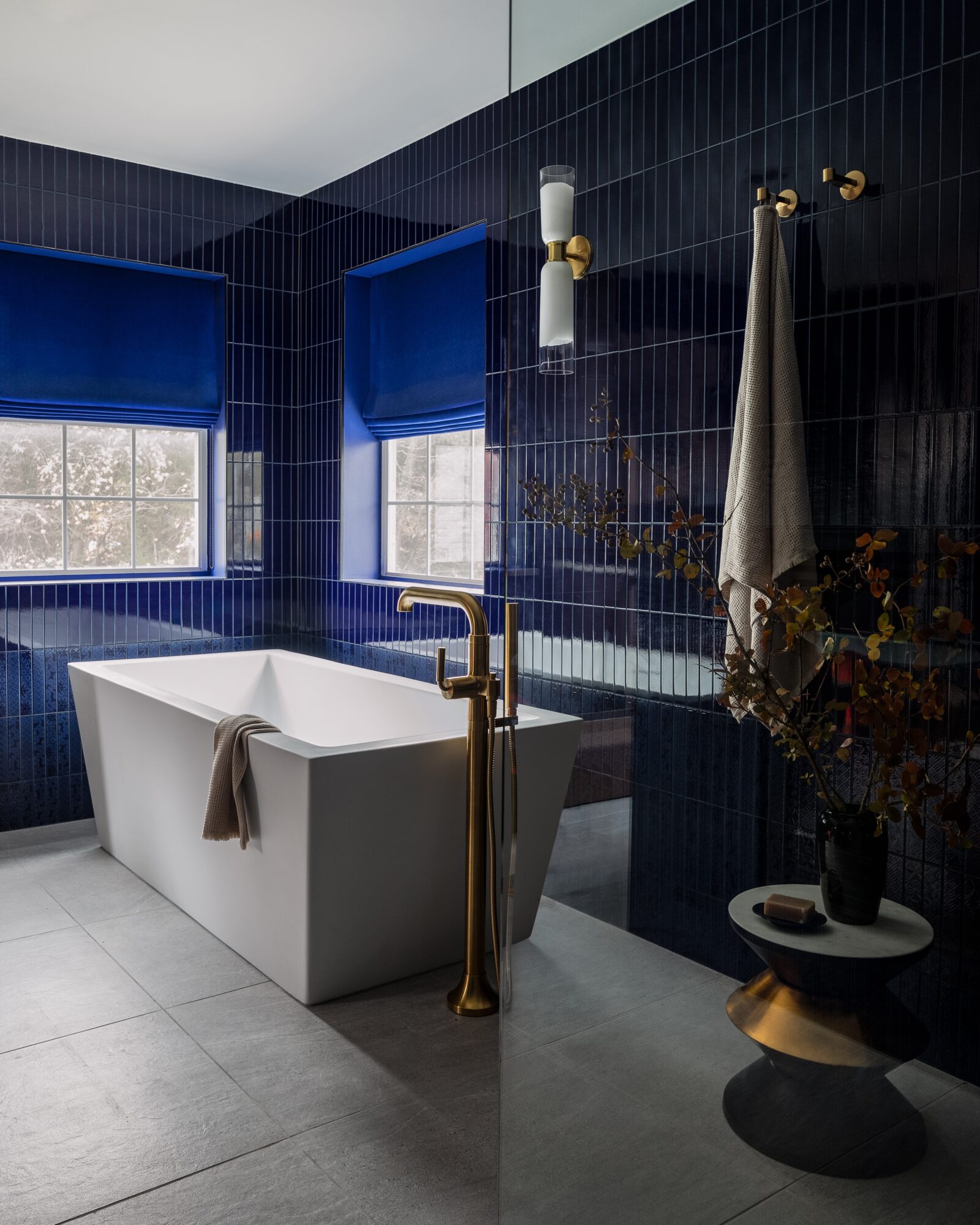 Modern bathroom with a white bathtub, blue window blinds, dark tiled walls, and a vase with flowers.