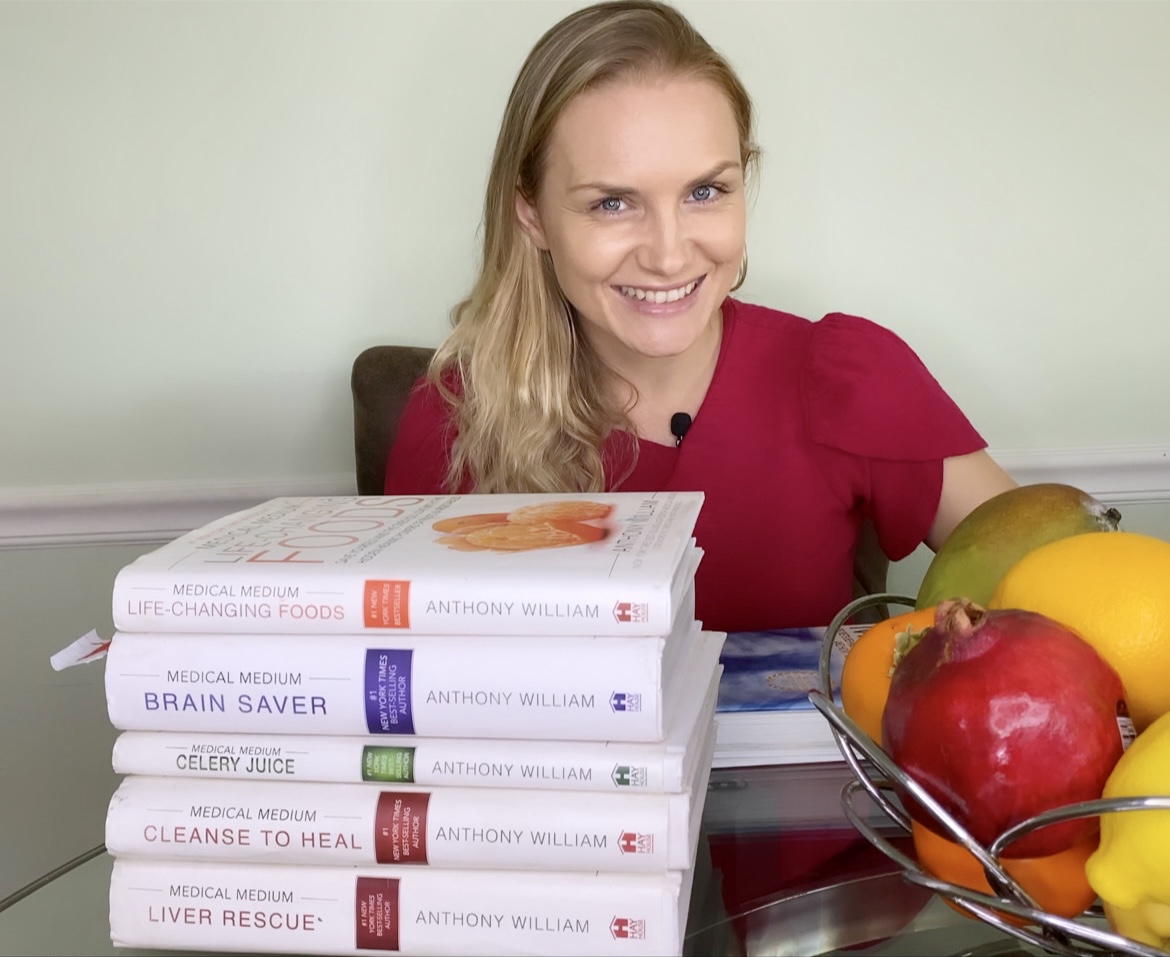 Woman with blonde hair smiling at camera, books, and a bowl of fruit on table, light green wall background.