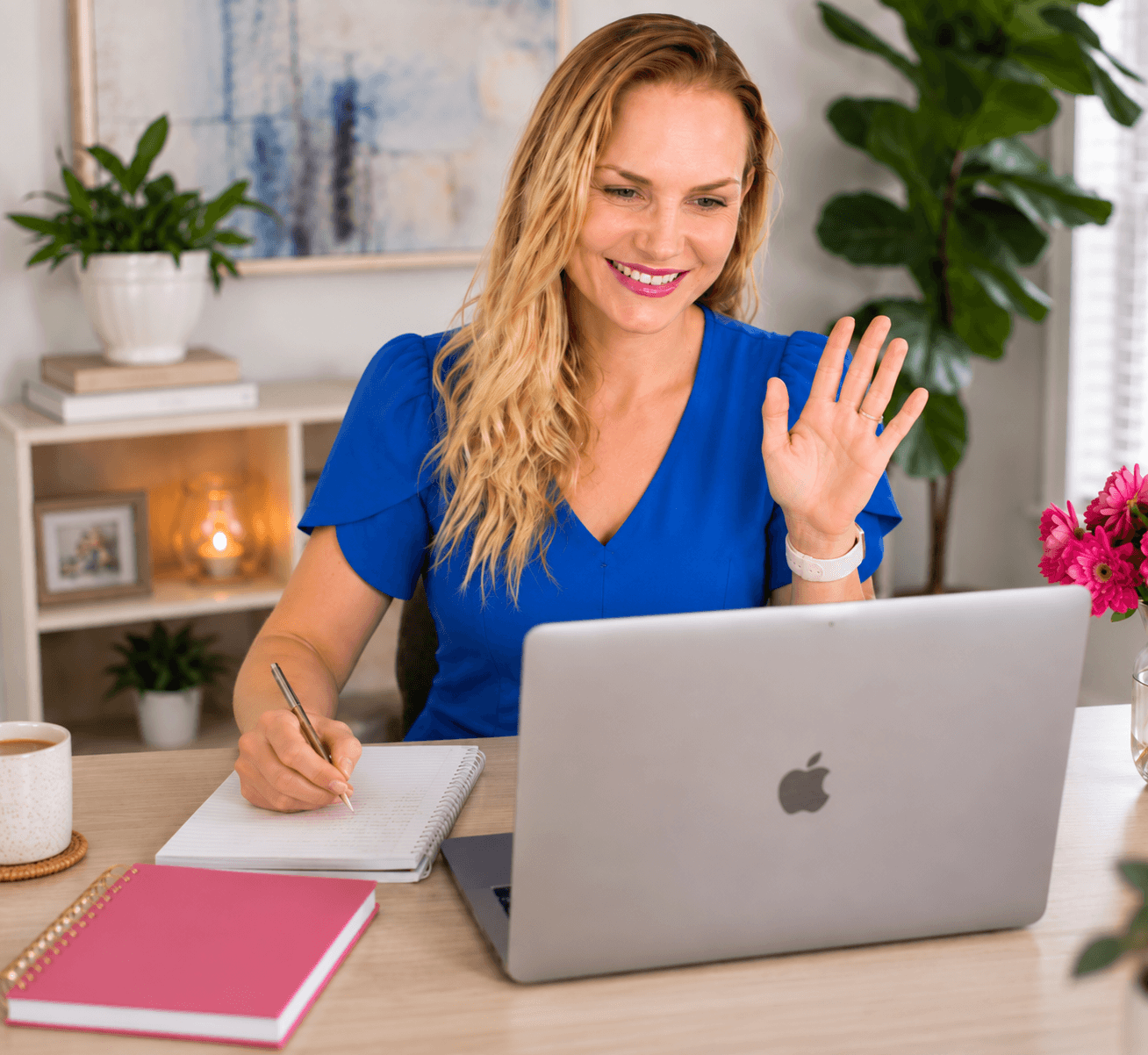 Woman in blue scrubs smiling and waving at a laptop, sitting at a desk with notebooks and plants nearby.