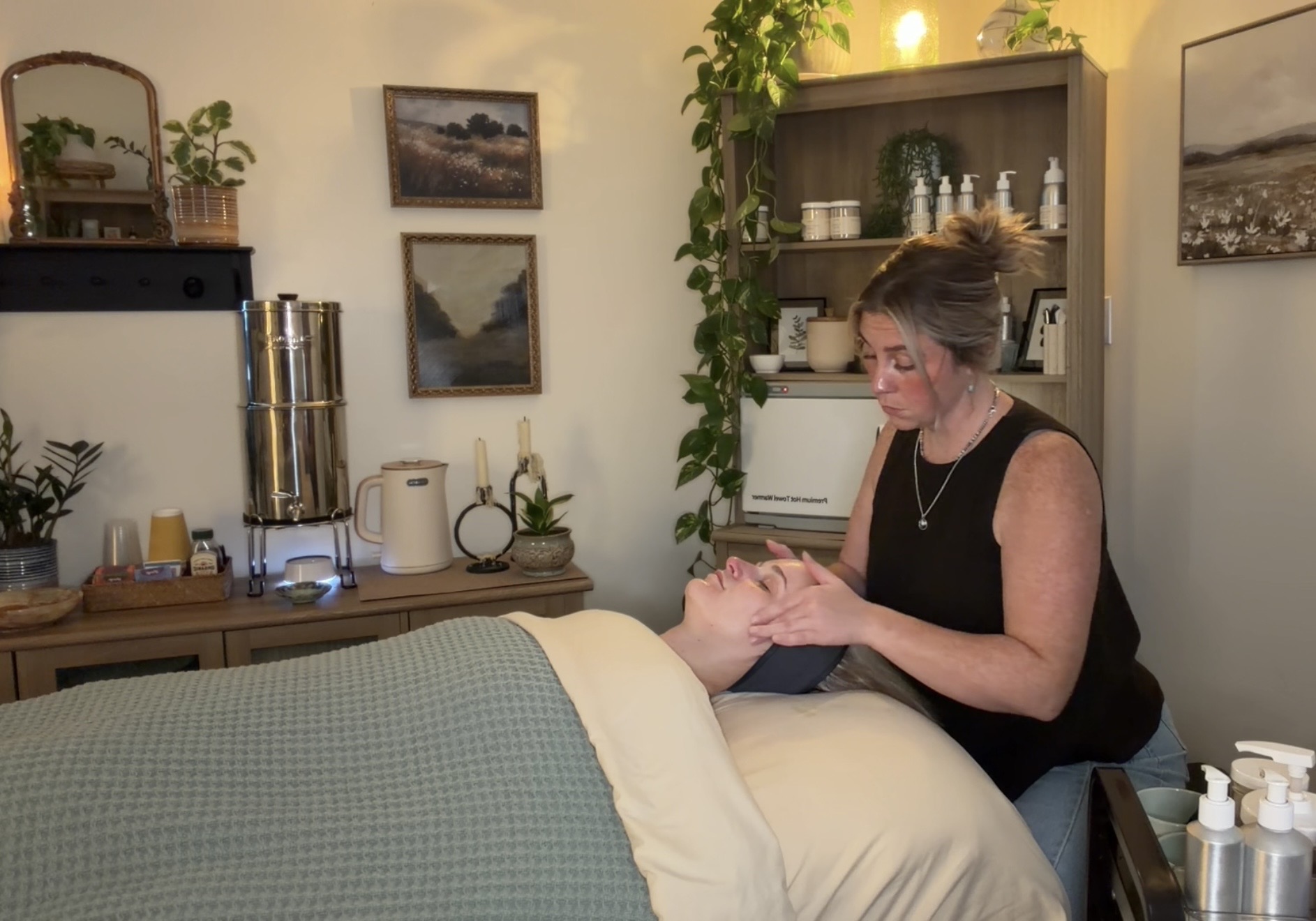 Woman giving a massage to a person lying on a massage table in a cozy room with shelves and framed pictures.