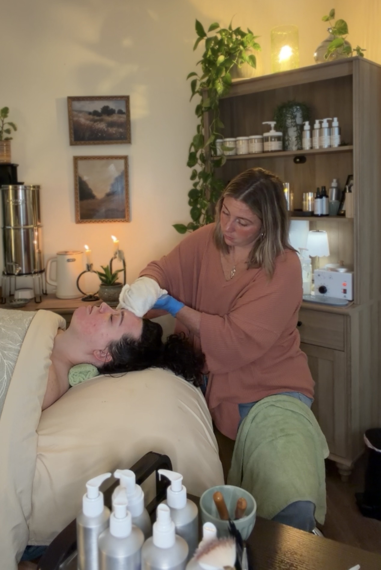 A woman receives a facial treatment from a professional in a cozy room with plants and shelves, warm lighting, and skincare products.