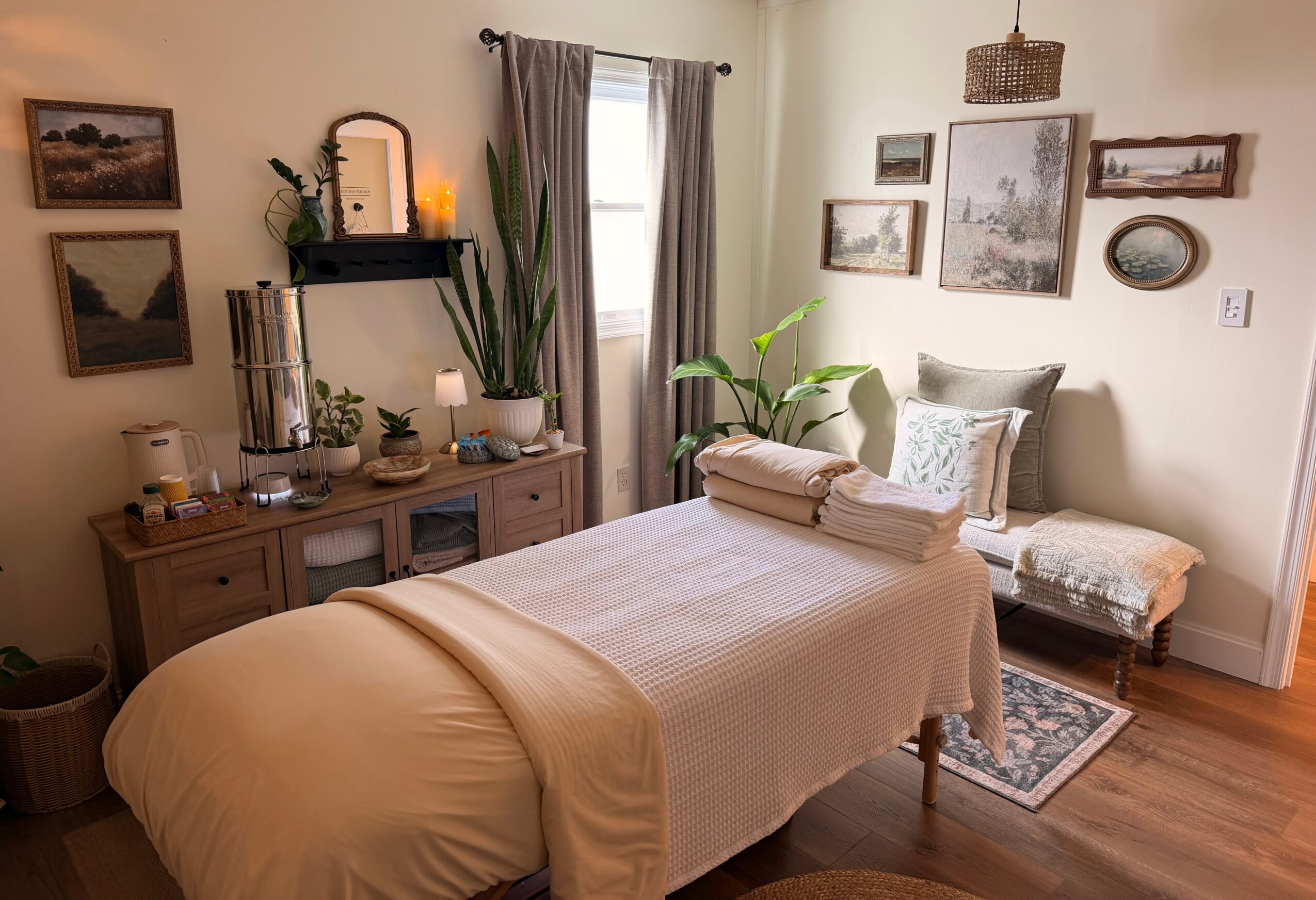 A cozy bedroom with a bed, side table, and wall art, featuring natural light from a window with curtains.