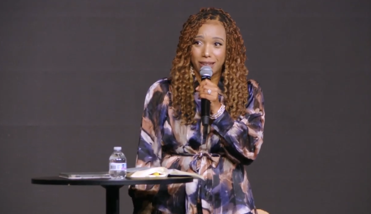 Woman with curly hair speaking into a microphone on stage, with a table holding a water bottle and papers nearby.