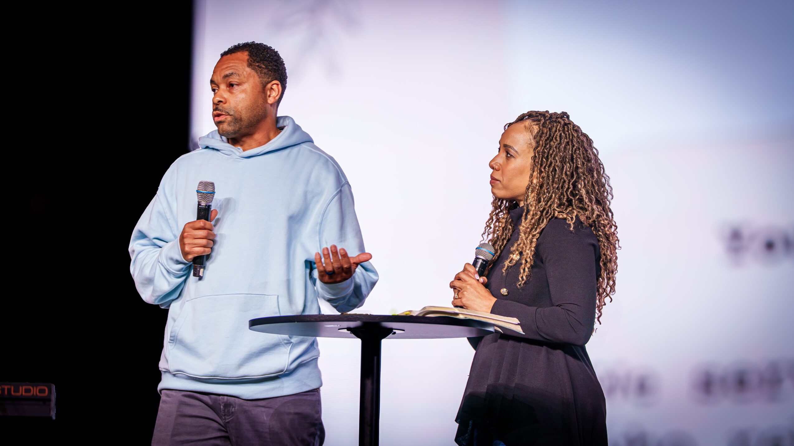 Two people stand on stage, one holding a microphone and speaking, the other listening attentively, with a plain background.