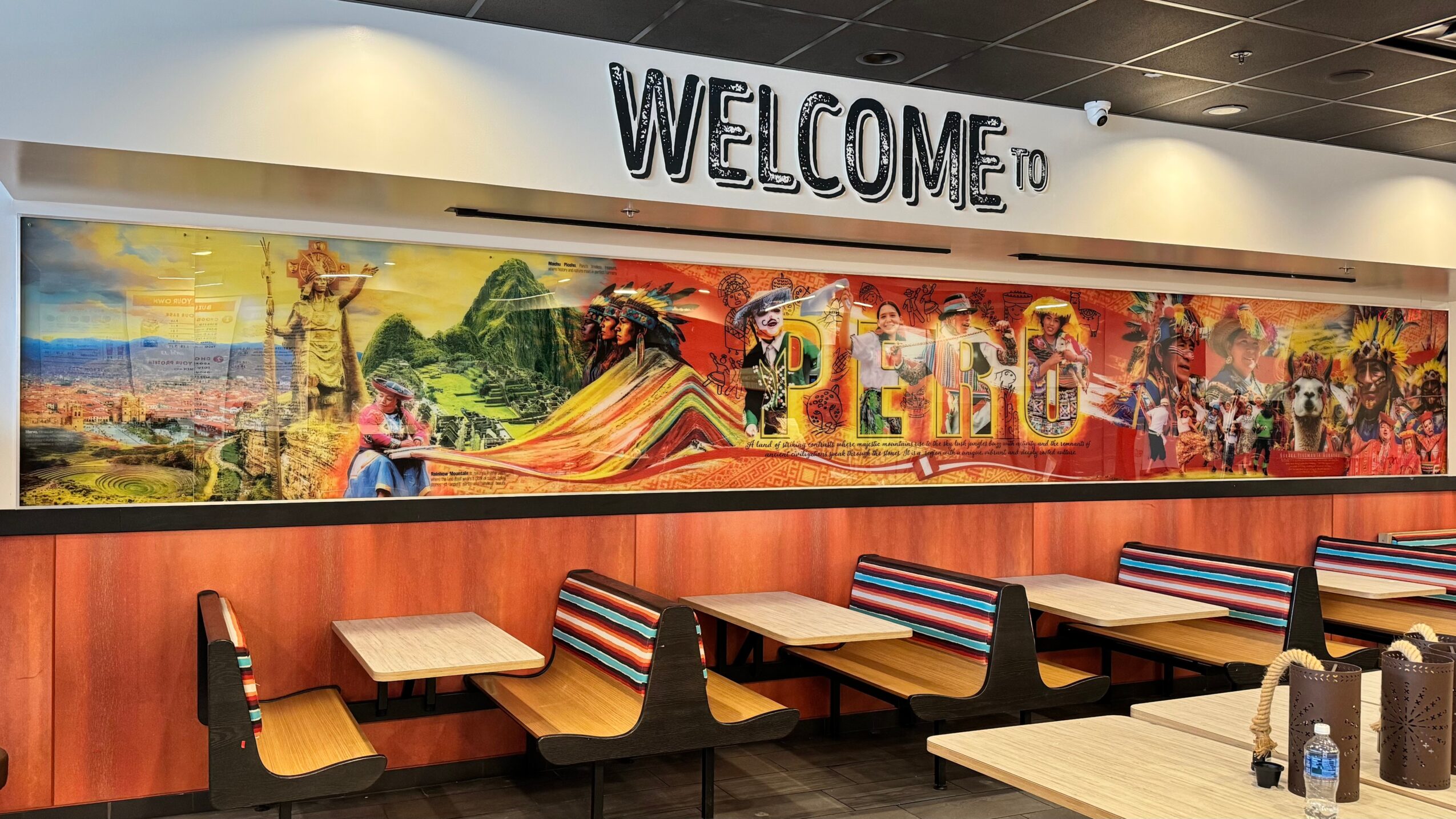 Welcome sign above colorful mural in a restaurant with tables and chairs, no people present.