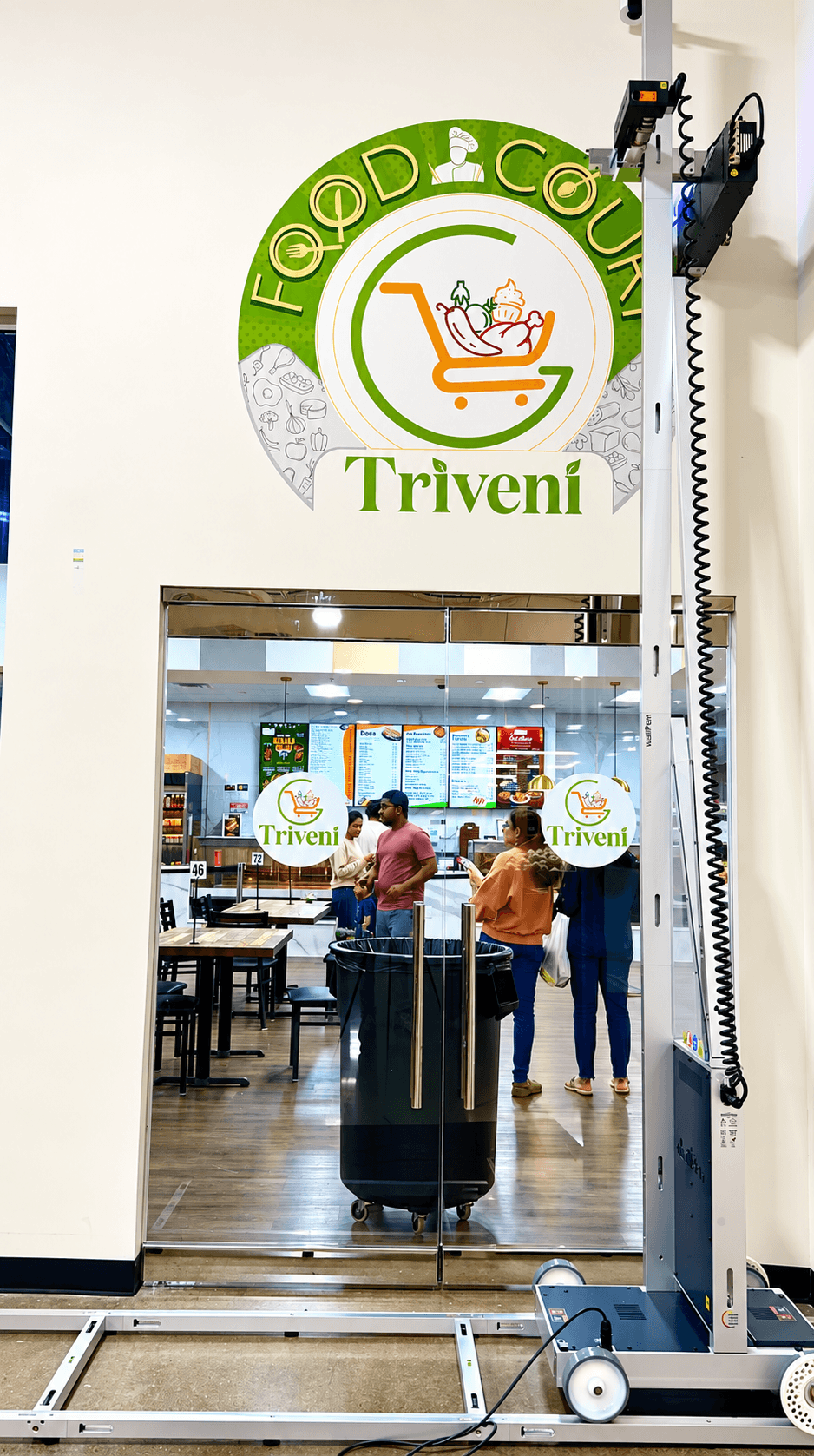 View of a grocery store entrance with people shopping inside, store logo with shopping cart and text 'Triveni', and a white wall.