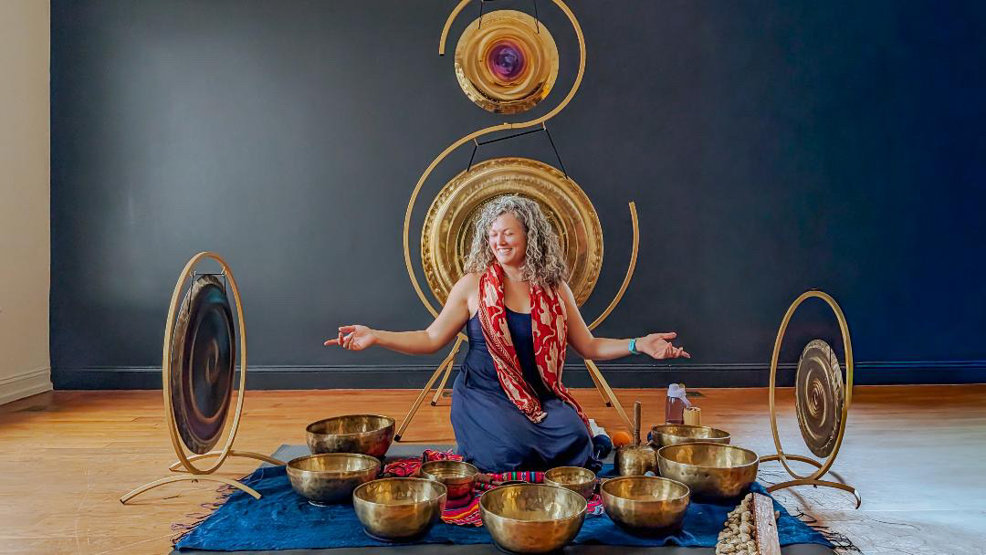 Woman sitting cross-legged on a blue rug with singing bowls around her, arms outstretched, in front of a dark wall with circular decorations.