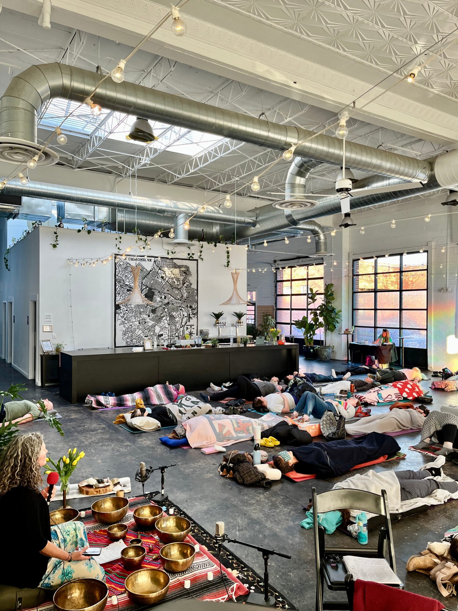 People lying on the floor in a spacious room with large windows, colorful decorations, and plants, participating in a group activity.
