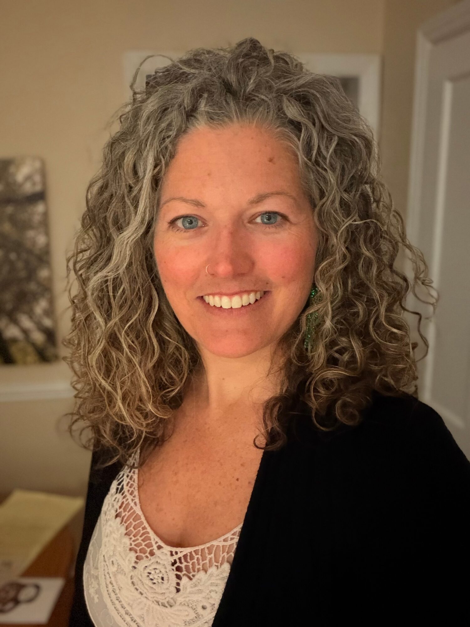 Woman with curly hair smiling indoors, wearing a white lace top and black cardigan.