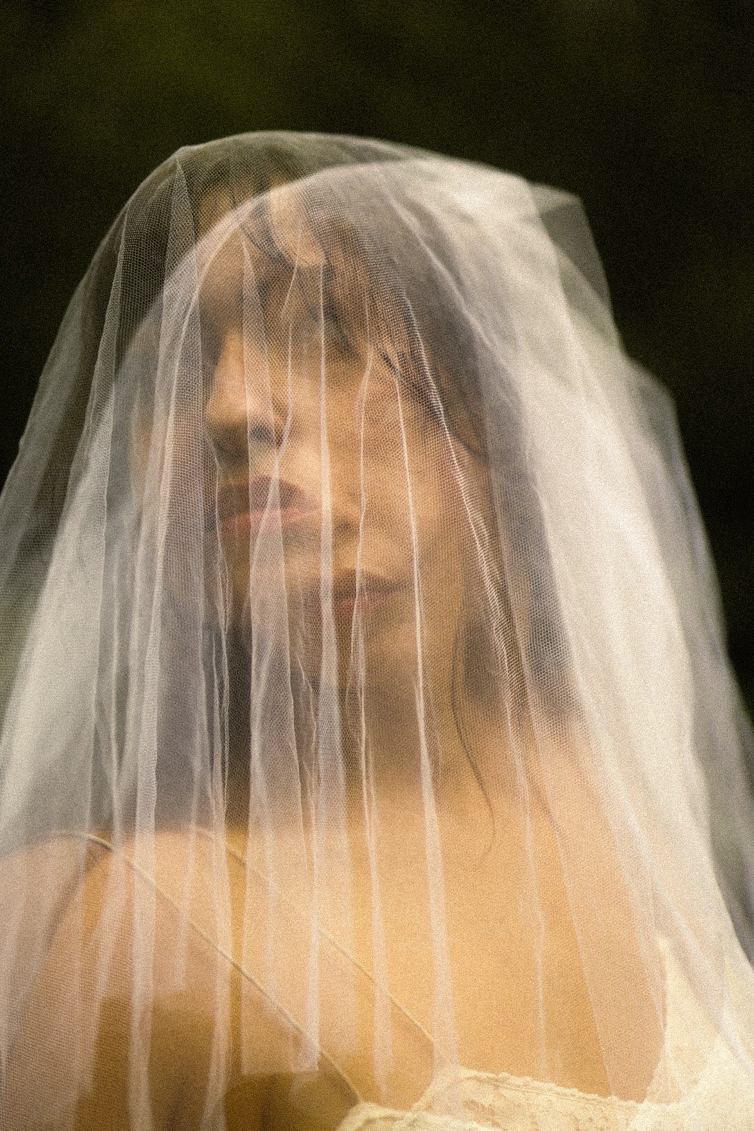 Woman with a veil covering her face, looking to the side, outdoors, dark background.