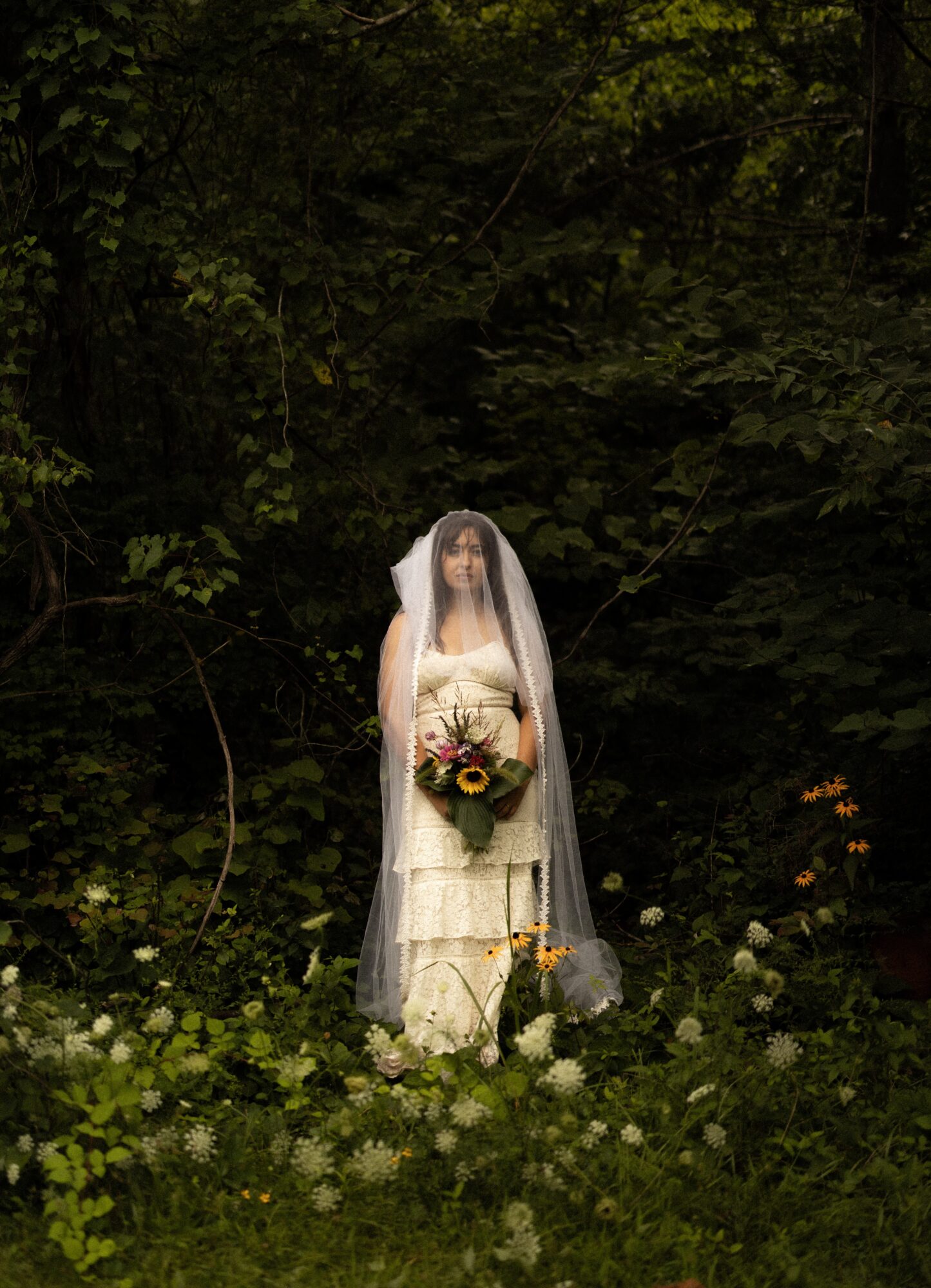 A person in a wedding dress holding a bouquet stands in a forest clearing with green foliage and white flowers.