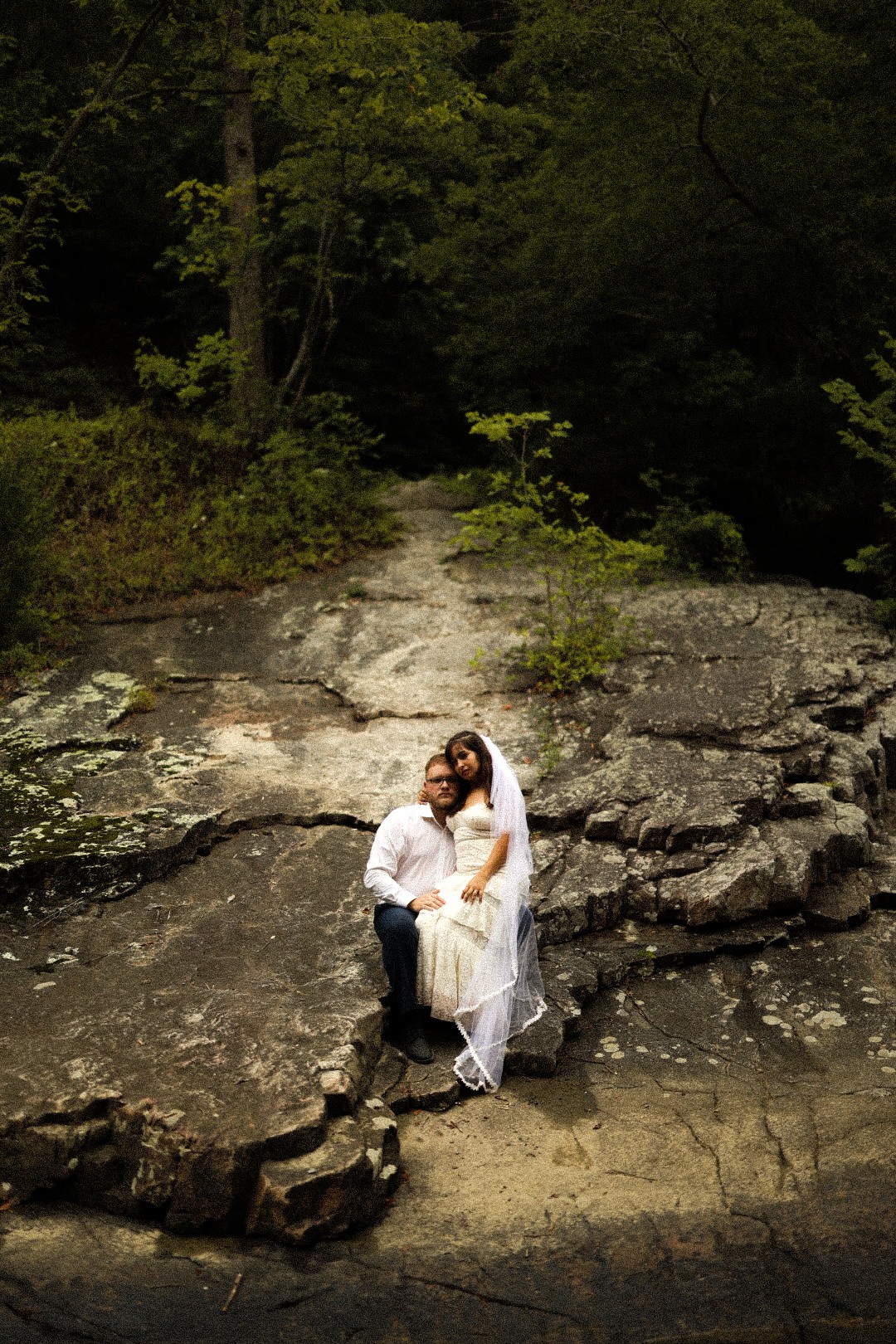 A couple sitting on rocks near a forested area, with trees and greenery in the background.