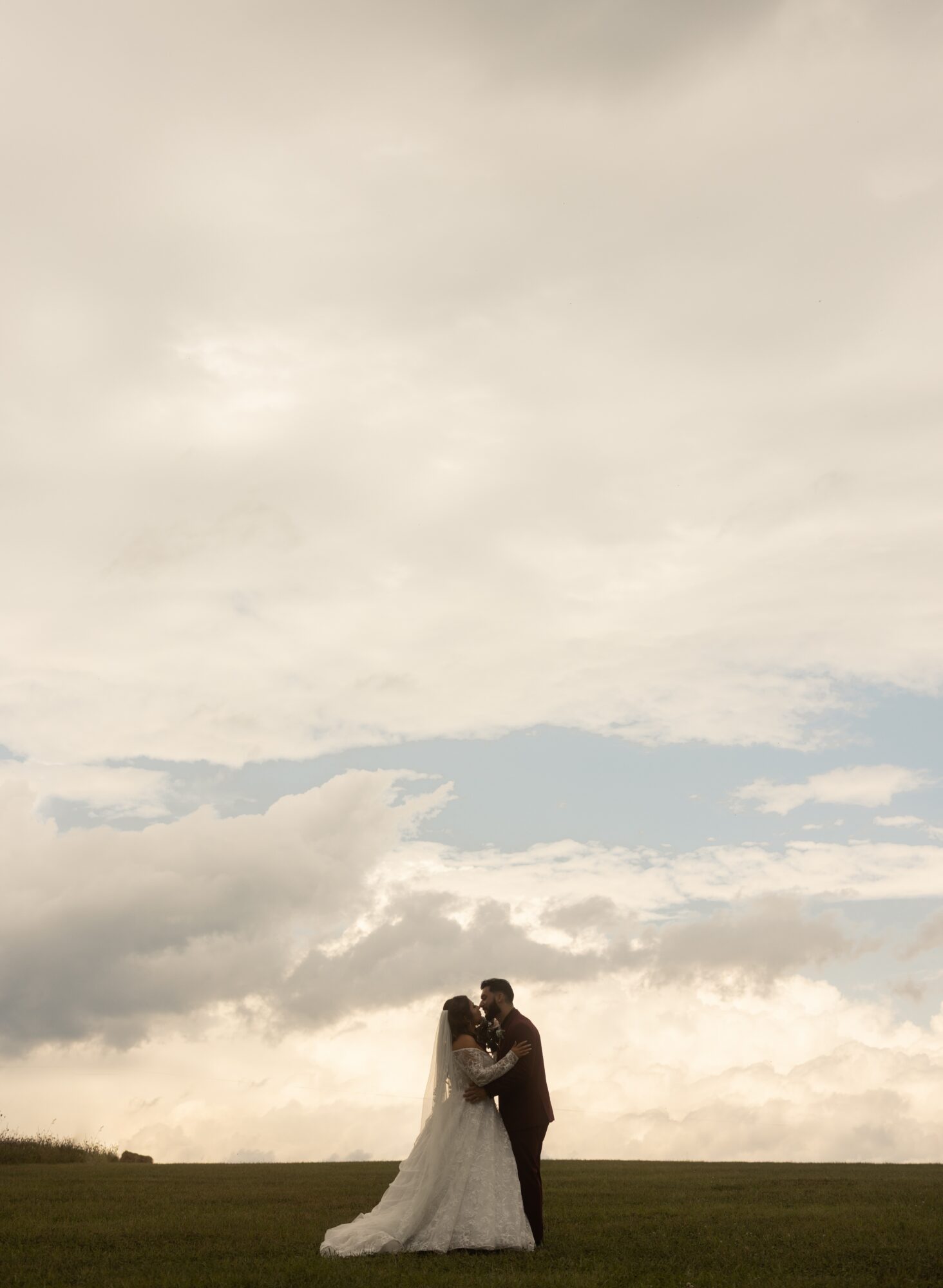 A couple stands together on a grassy field under a cloudy sky, embracing each other.
