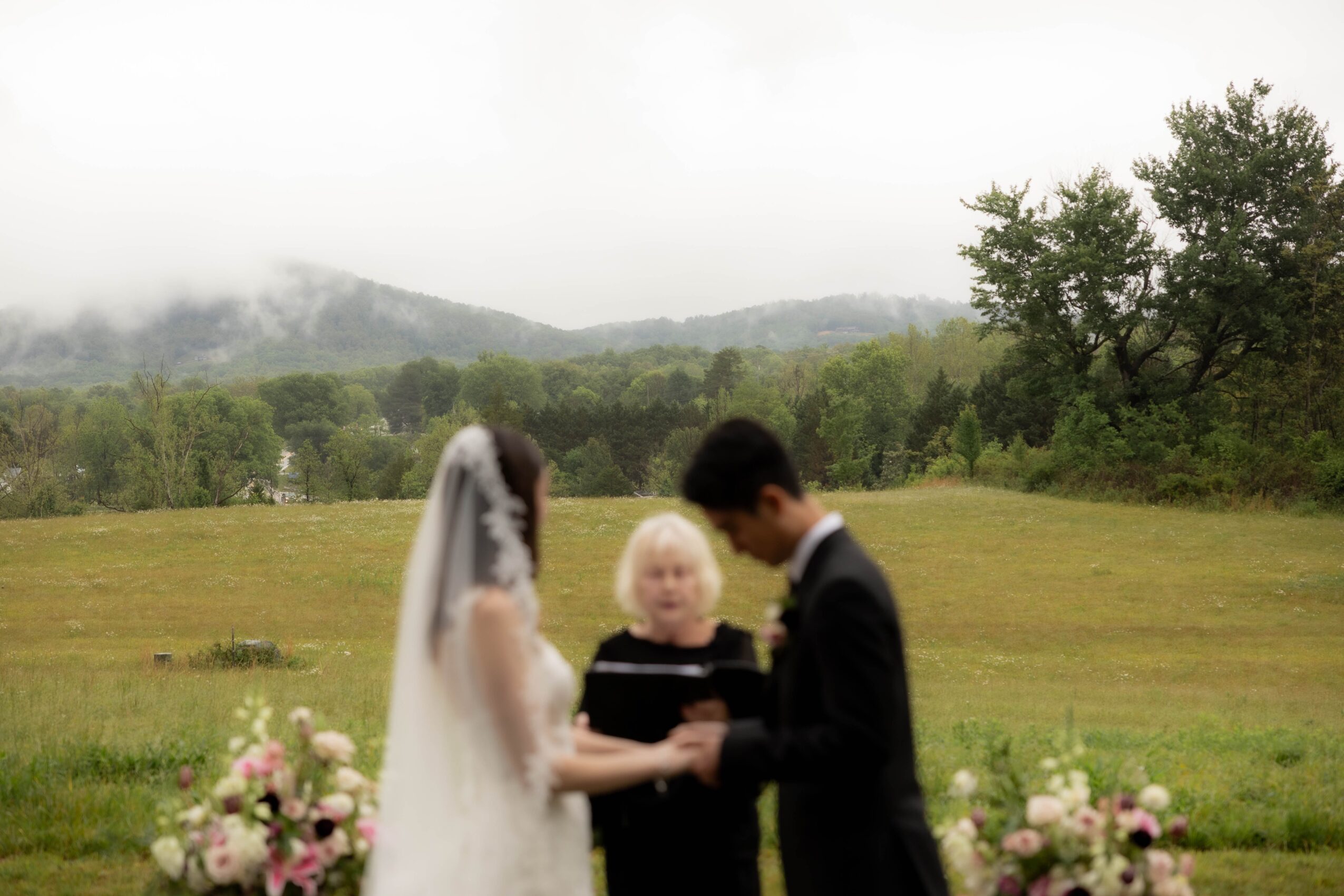 Three people, a bride, a groom, and a child, holding hands outdoors with a grassy field and trees in the background.